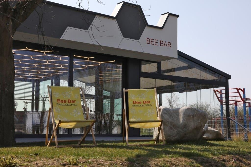Zwei gelbe Liegestühle vor der Bee Bar mit Glasfassade und geometrischem Dach.