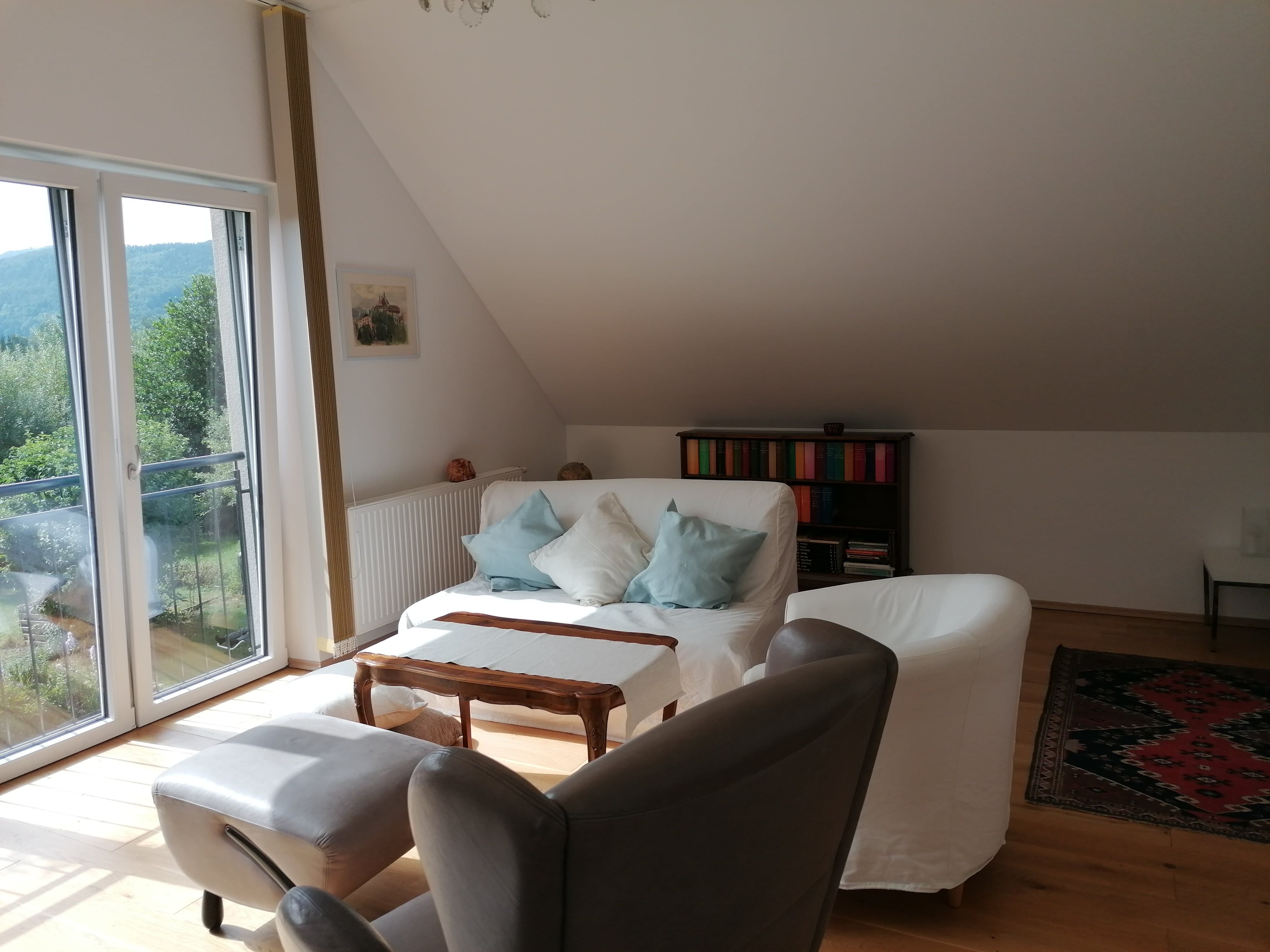 Heller Wohnbereich mit Sofa, Sesseln, Bücherregal und großem Fenster mit Blick ins Grüne.