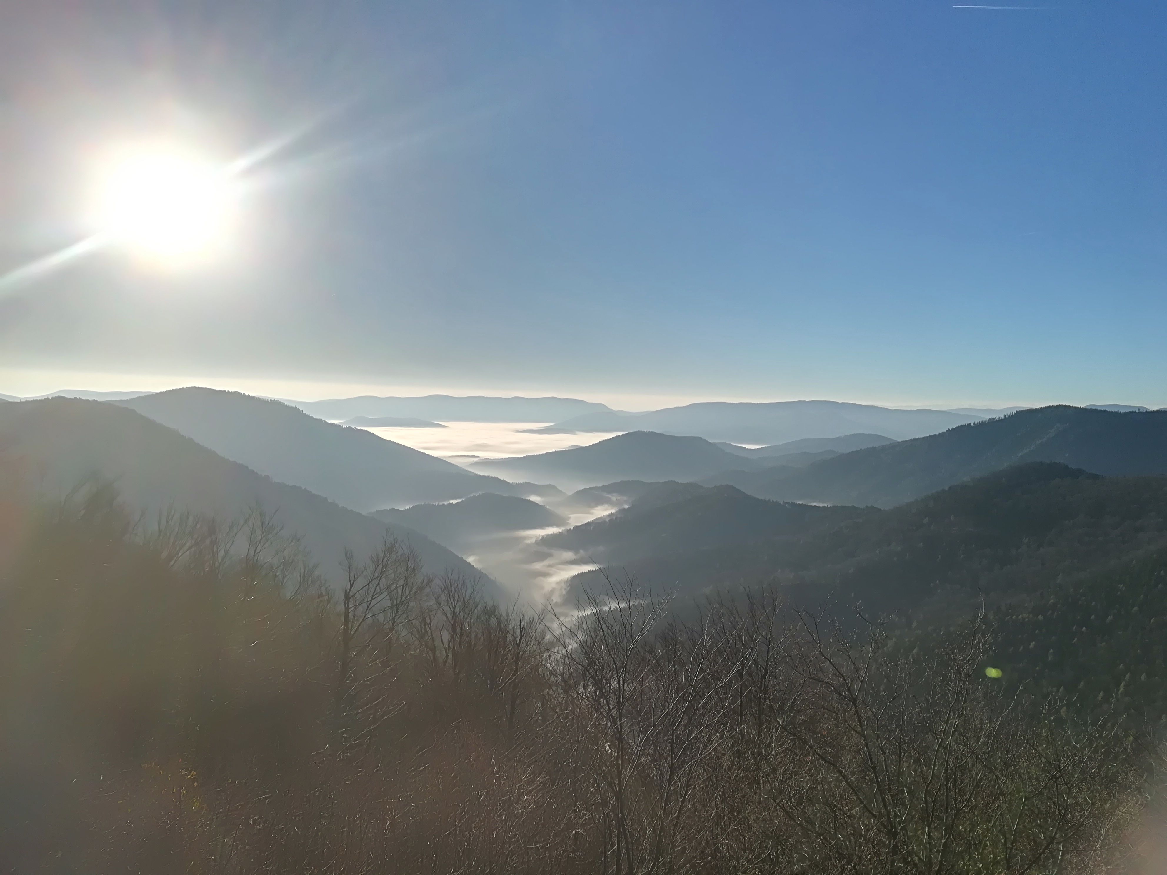 Blick von einem Berg auf nebelverhangene Täler und Hügel unter strahlend blauem Himmel mit Sonnenschein.