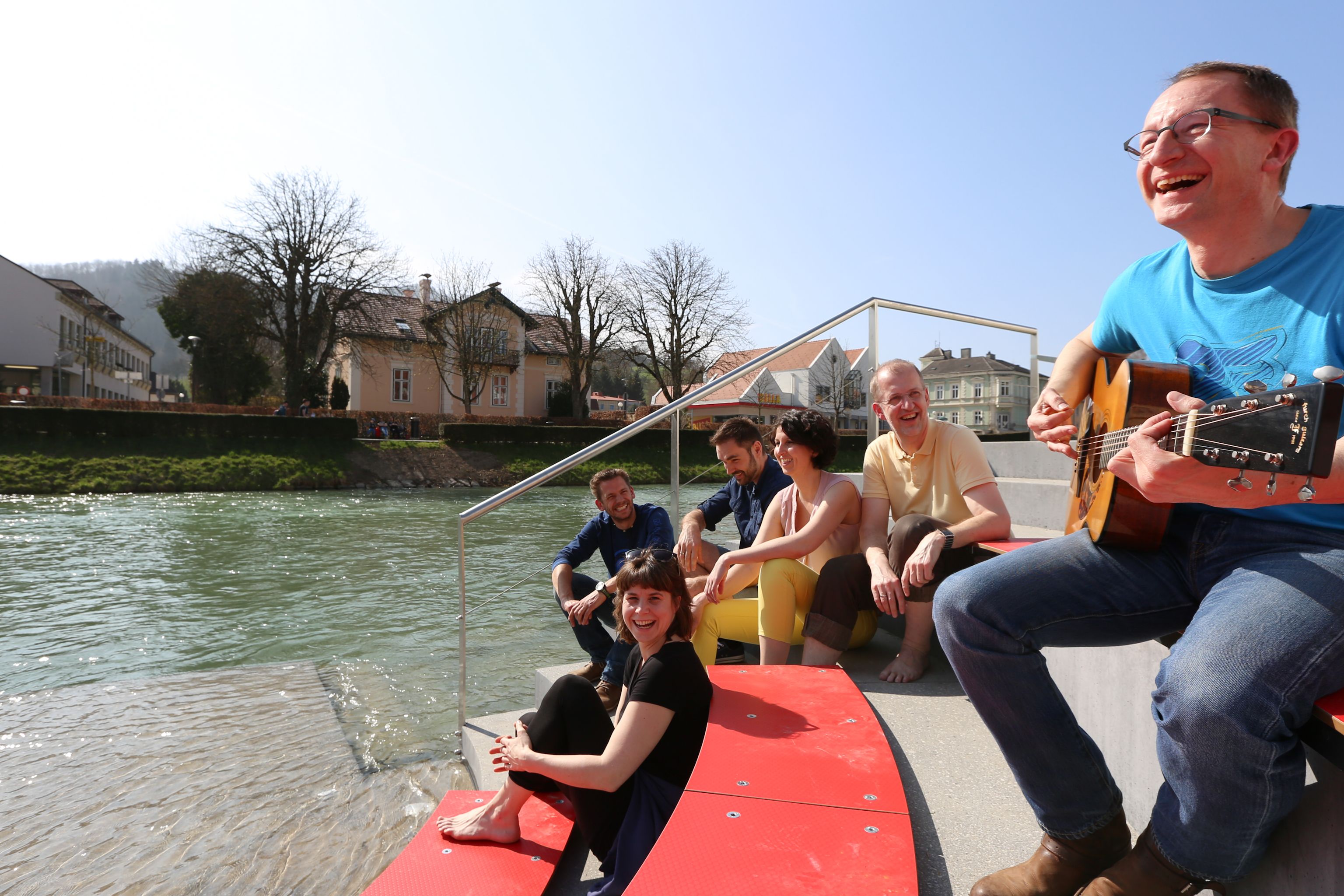Gruppe von Menschen sitzt lachend an einem Flussufer, eine Person spielt Gitarre.