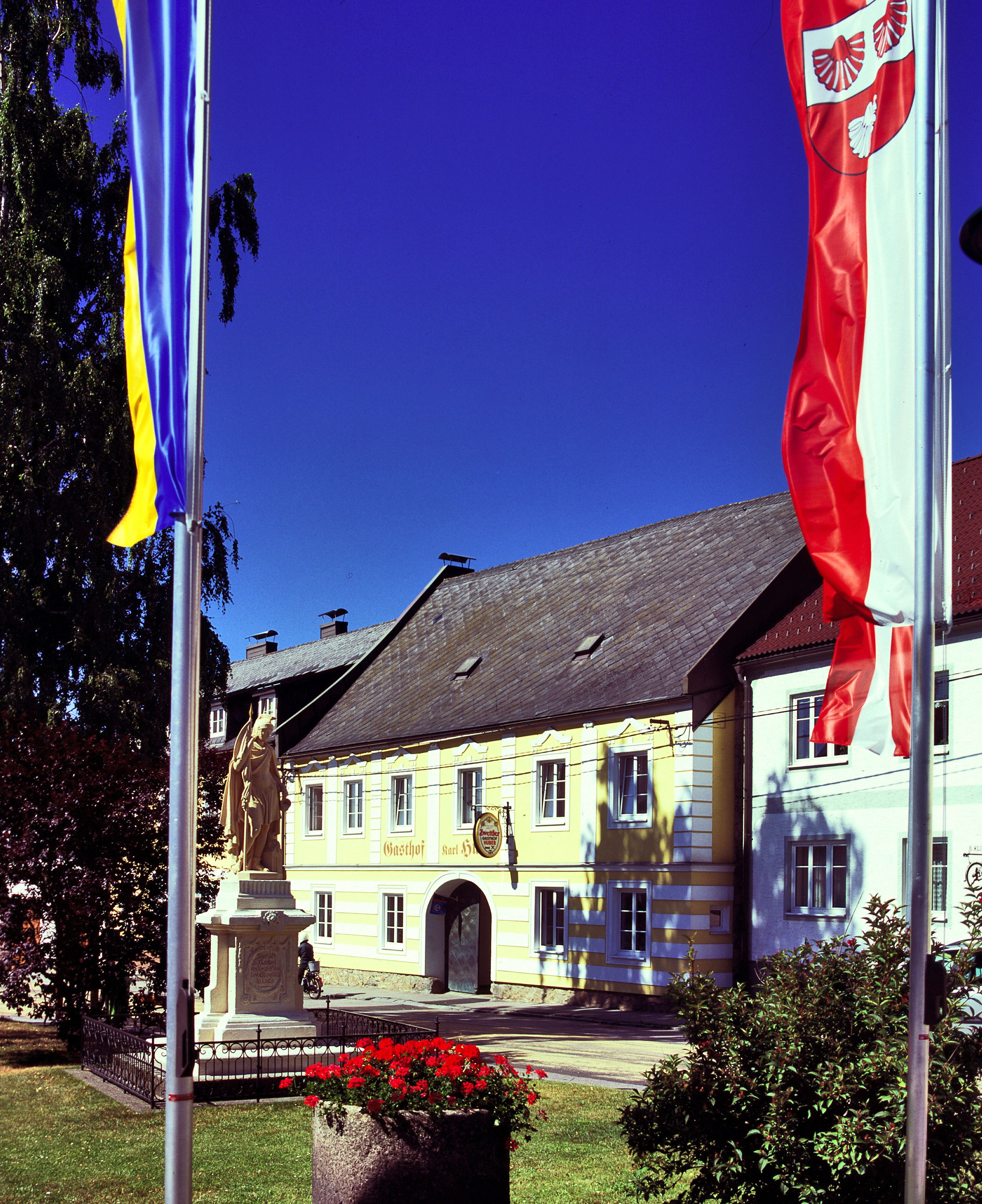 Gelbes Gebäude mit der Aufschrift 'Gasthof Karl Huber', umgeben von Fahnen und einer Statue.