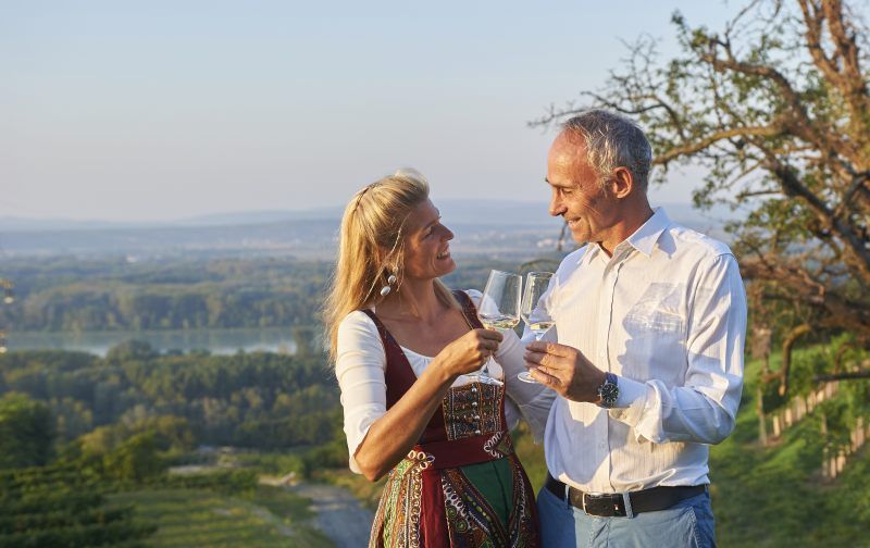 Ein Paar stößt mit Weingläsern in einem Weinberg an, im Hintergrund eine malerische Landschaft.