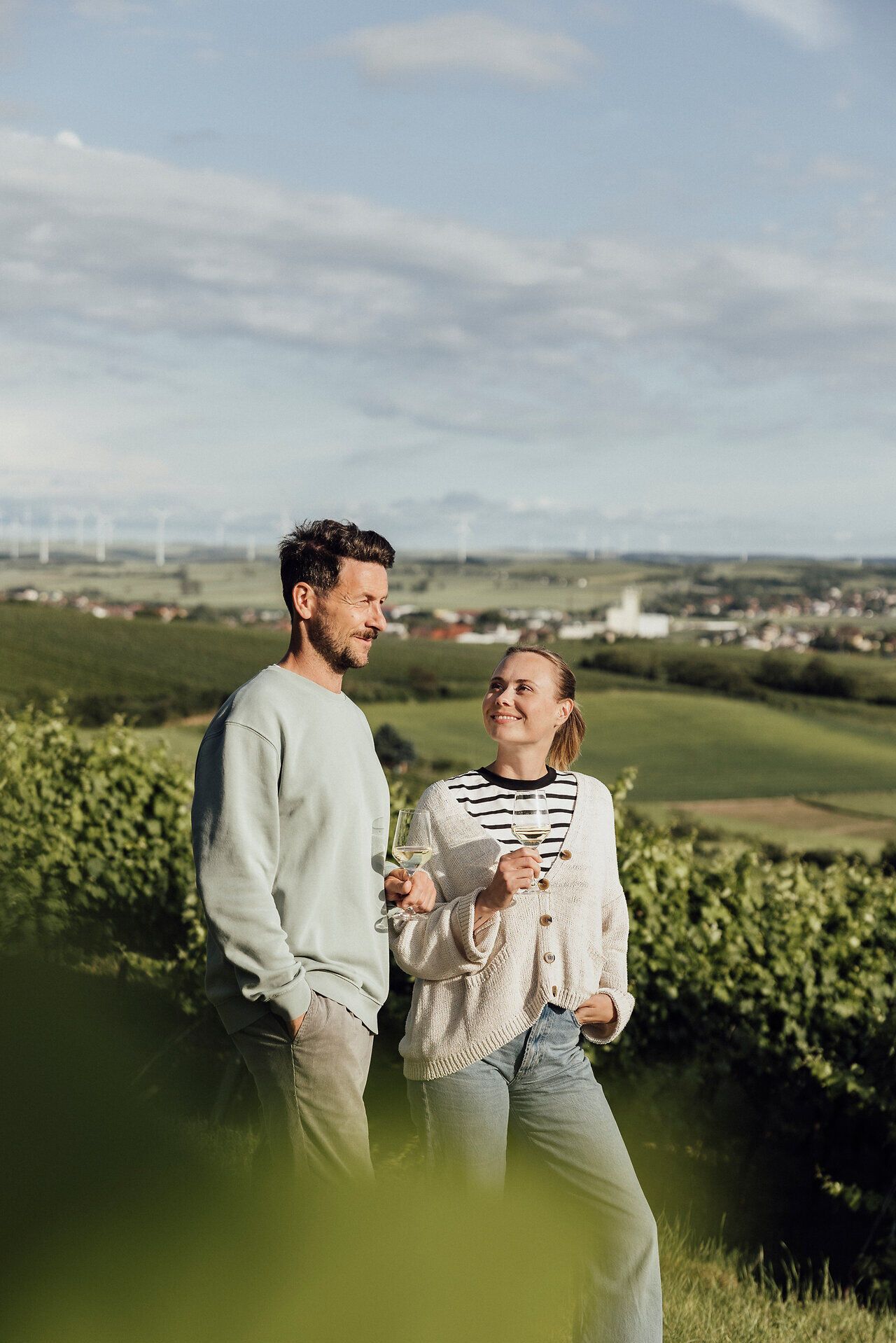 Inmitten der sanften Hügel des Weinviertels genießen zwei Menschen ein Glas Wein und lassen sich von der malerischen Landschaft verzaubern. Die grüne Umgebung und der weite Blick laden dazu ein, die Seele baumeln zu lassen und den Moment in vollen Zügen auszukosten.