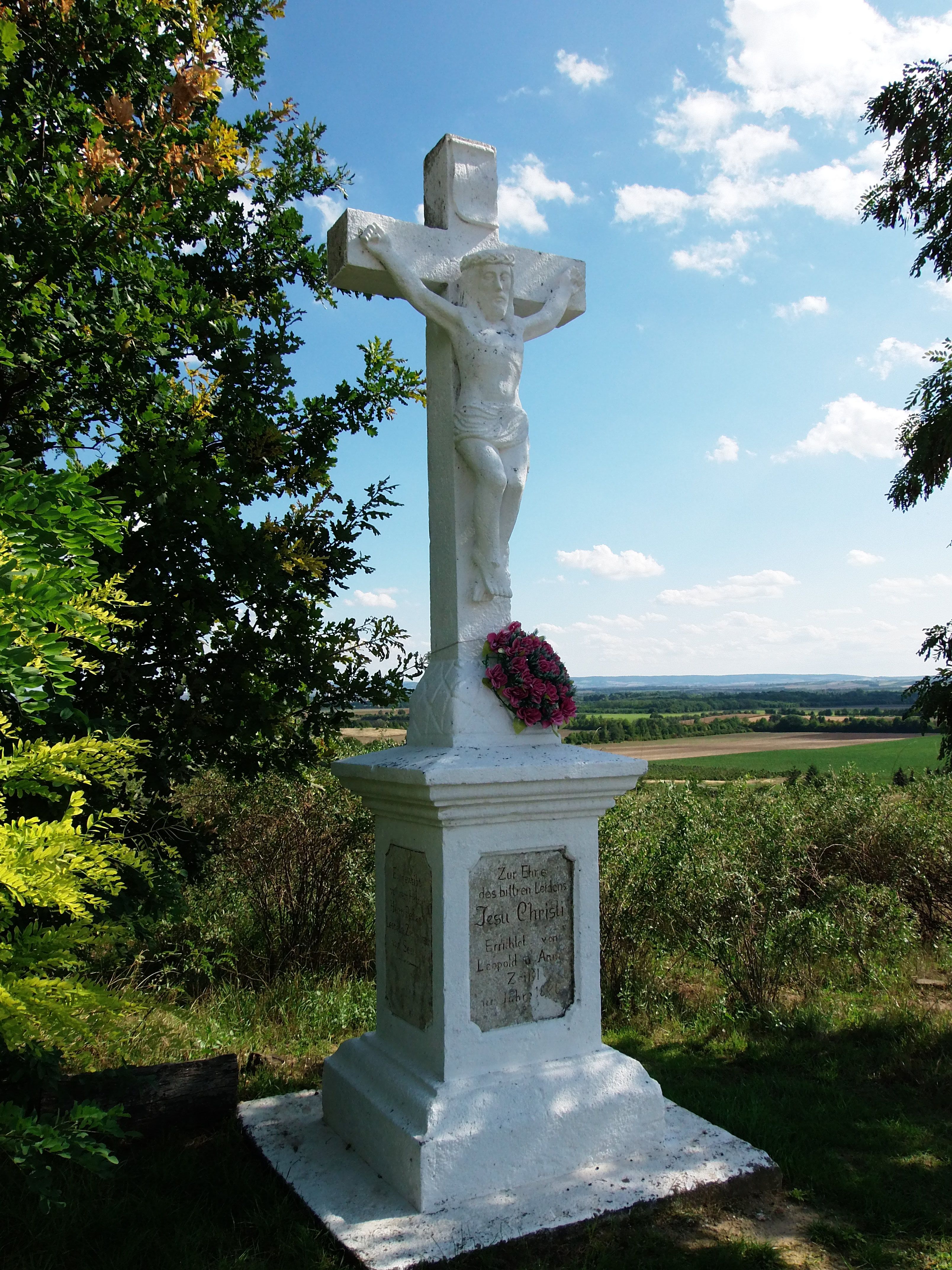 Weißes Steinkreuz mit Jesusfigur und Blumenkranz, umgeben von Bäumen und Landschaft im Hintergrund.