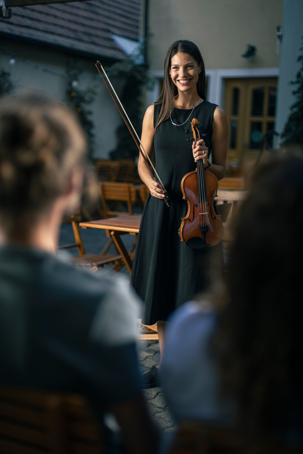 Geigerin in schwarzem Kleid steht mit Violine und Bogen im Innenhof eines Weinguts und lächelt dem sitzenden Publikum entgegen; im Hintergrund Holztische und Weinflaschen, stimmungsvolle Atmosphäre bei einer Kulturveranstaltung im Weingut.