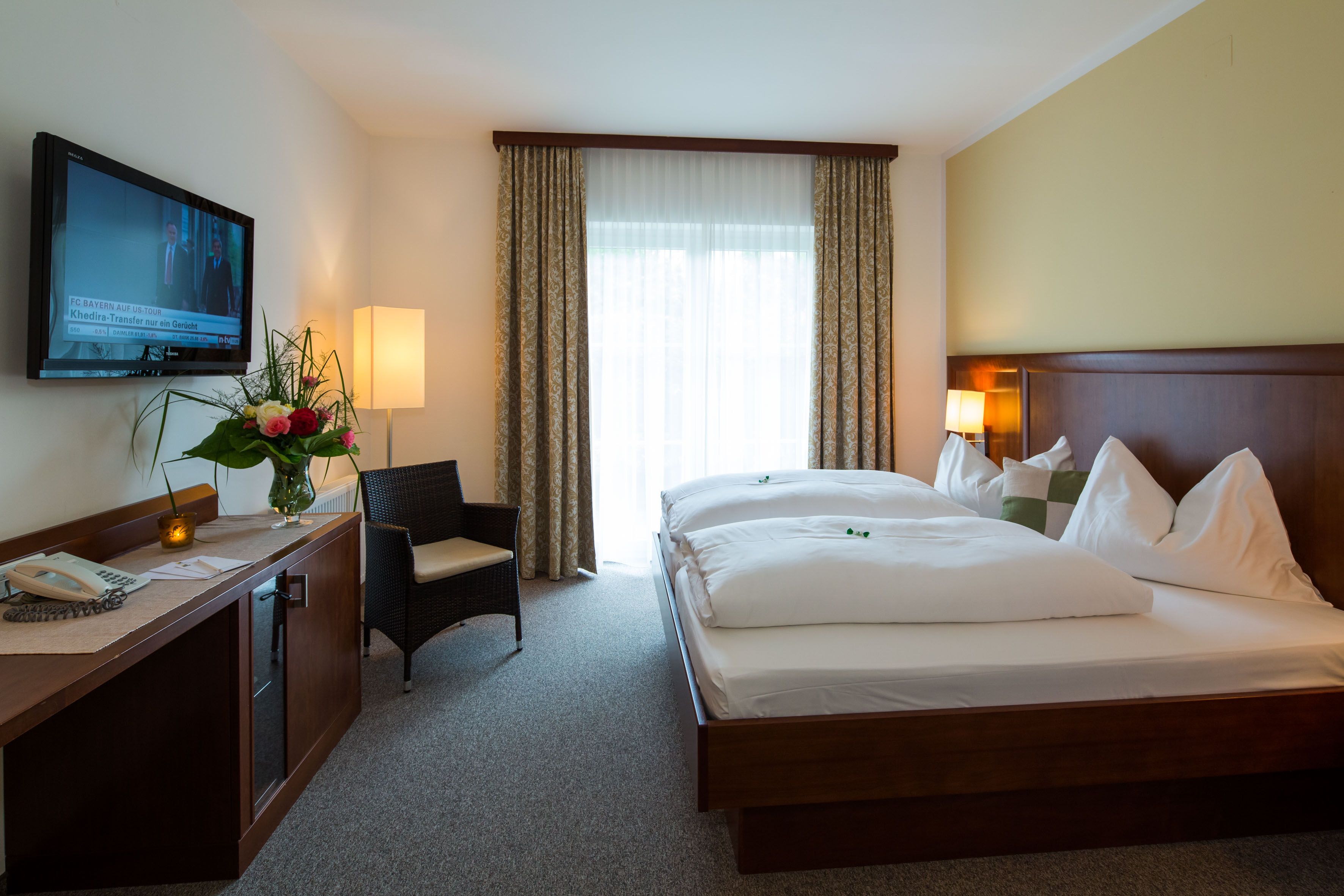 Ein Hotelzimmer mit Doppelbett, Fernseher, Sessel und Blumen auf einem Tisch.