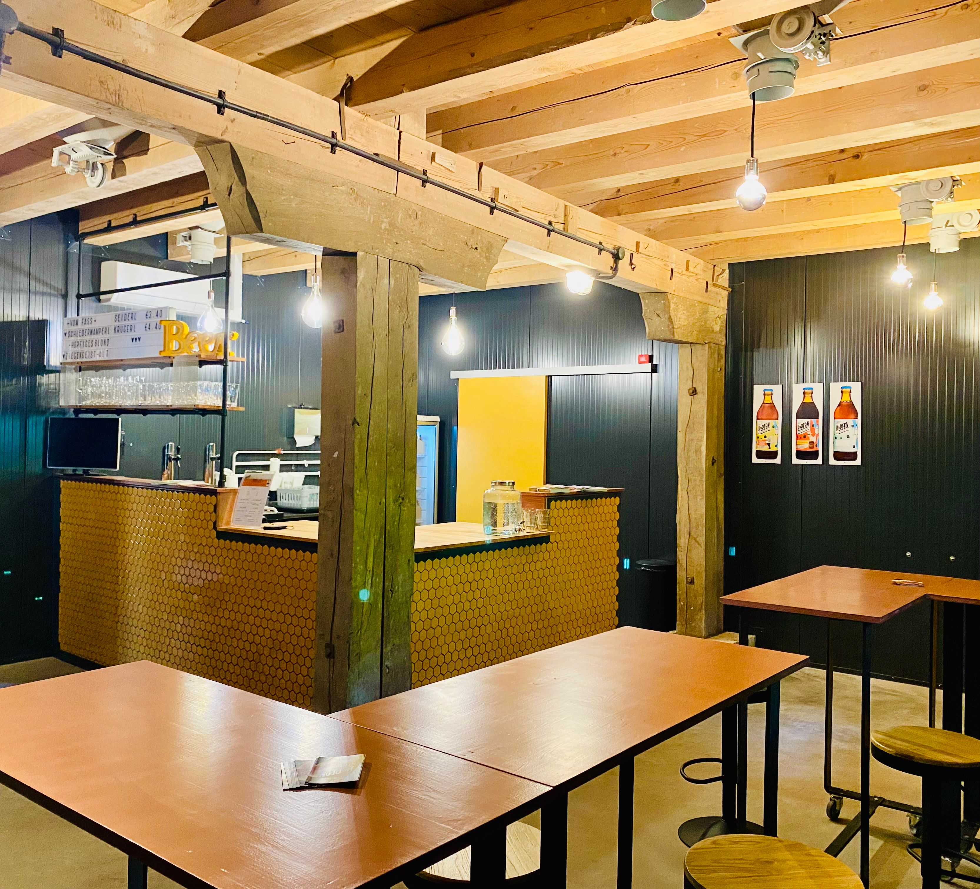 Innenansicht eines modernen Cafés mit Holzbalken, Bartresen und hohen Tischen.