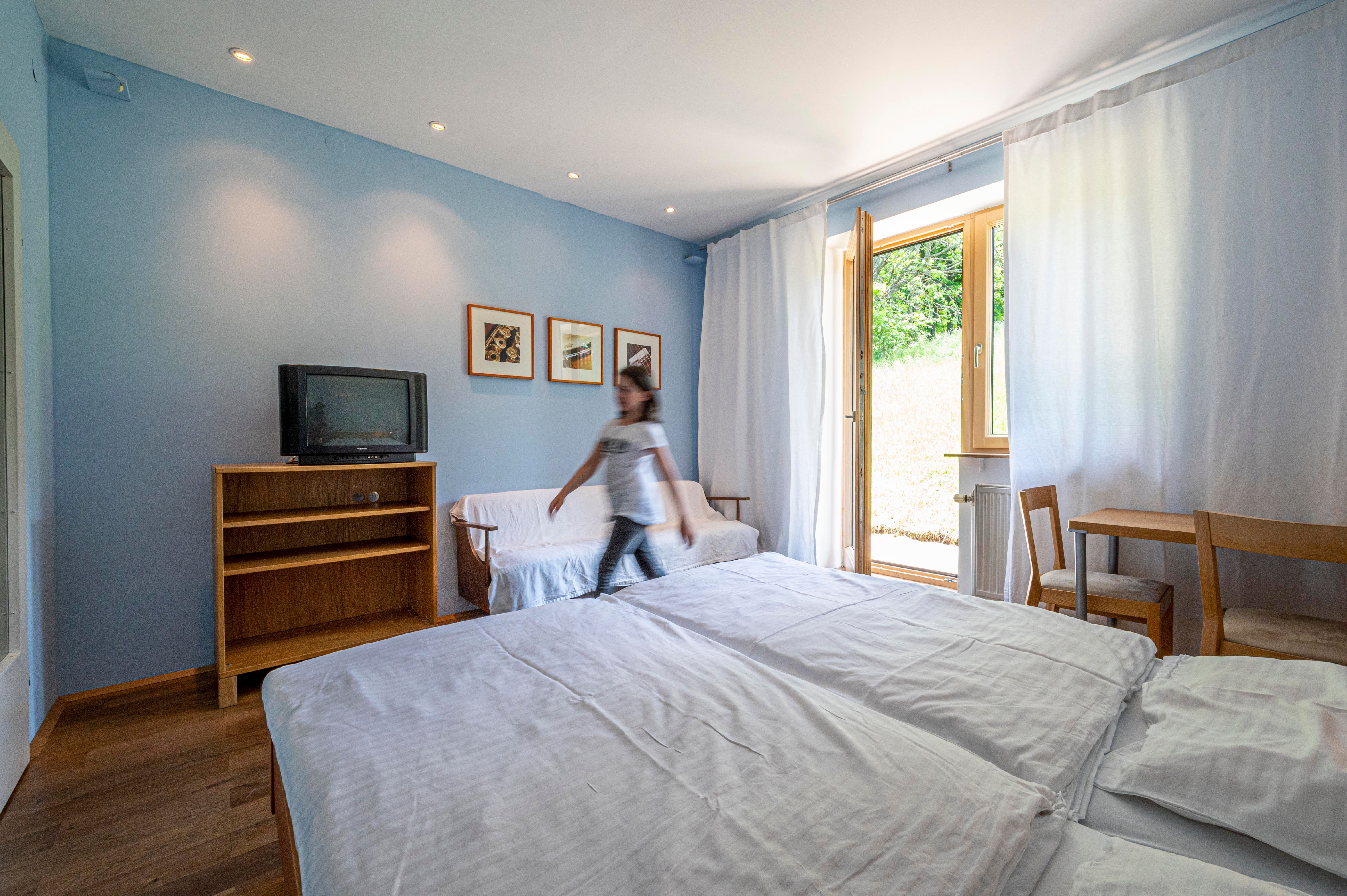 Ein helles Zimmer mit blauen Wänden, einem Bett, einem Fernseher und einem kleinen Tisch. Eine Person geht durch den Raum.