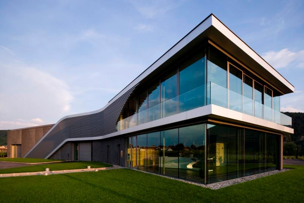 Modernes Gebäude mit Glasfassade und geschwungenem Dach, umgeben von grüner Wiese.