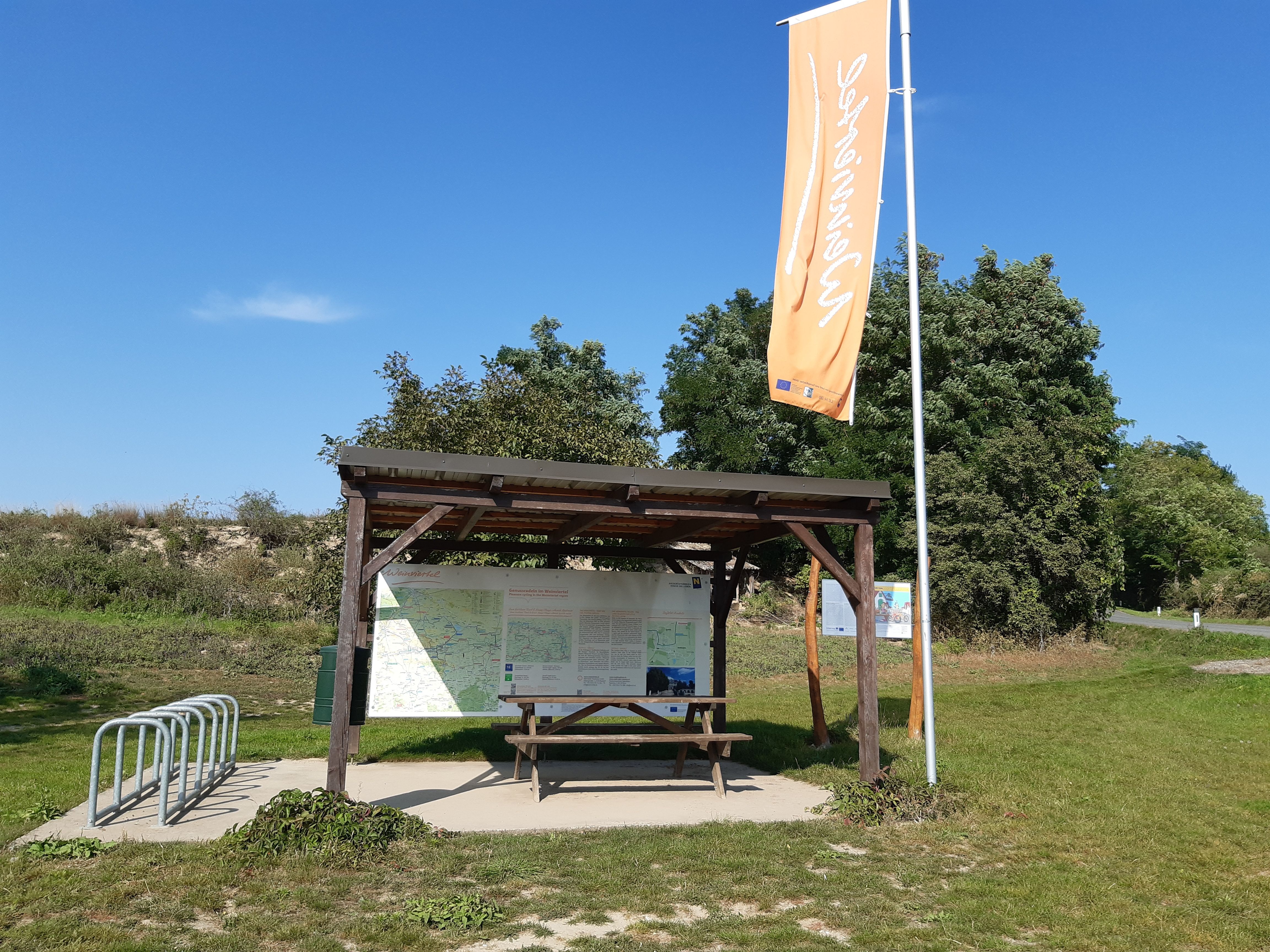 Raststation mit überdachtem Tisch, Fahrradständer und Infotafeln im Grünen.