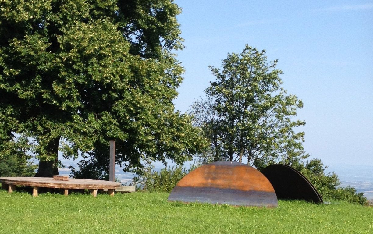 Holzplattform und Metallskulpturen auf einer Wiese mit Bäumen im Hintergrund.