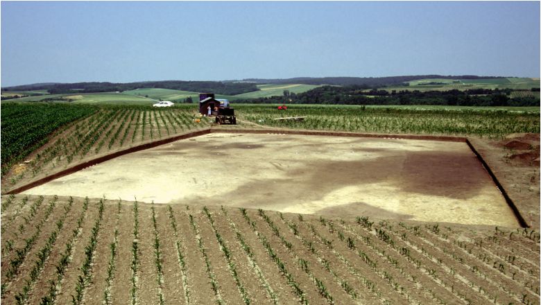 Archäologische Ausgrabungsstätte auf einem Feld mit landwirtschaftlichen Maschinen im Hintergrund.