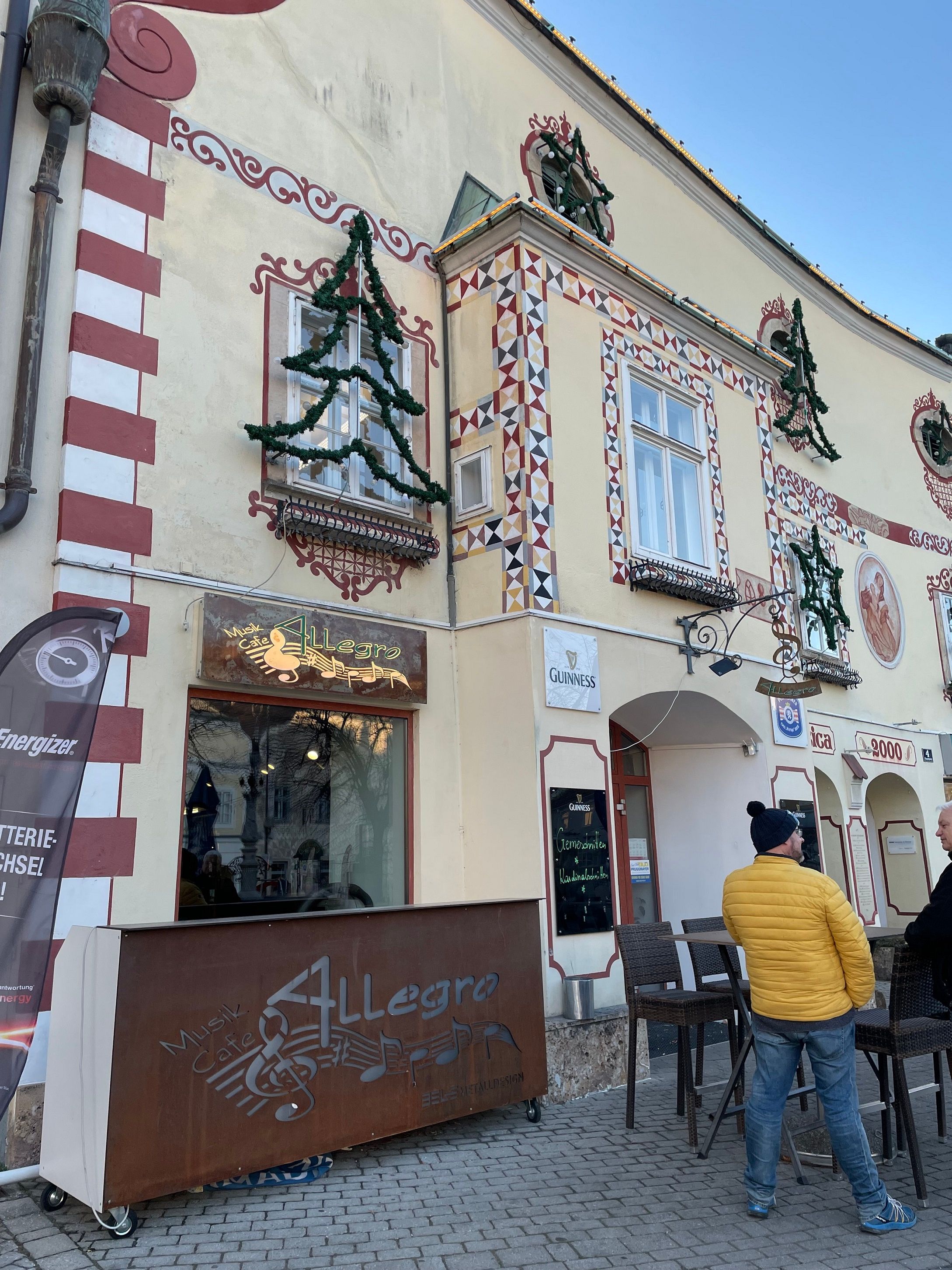 Außenansicht des Cafe Allegro mit dekorierter Fassade und zwei Personen im Gespräch.