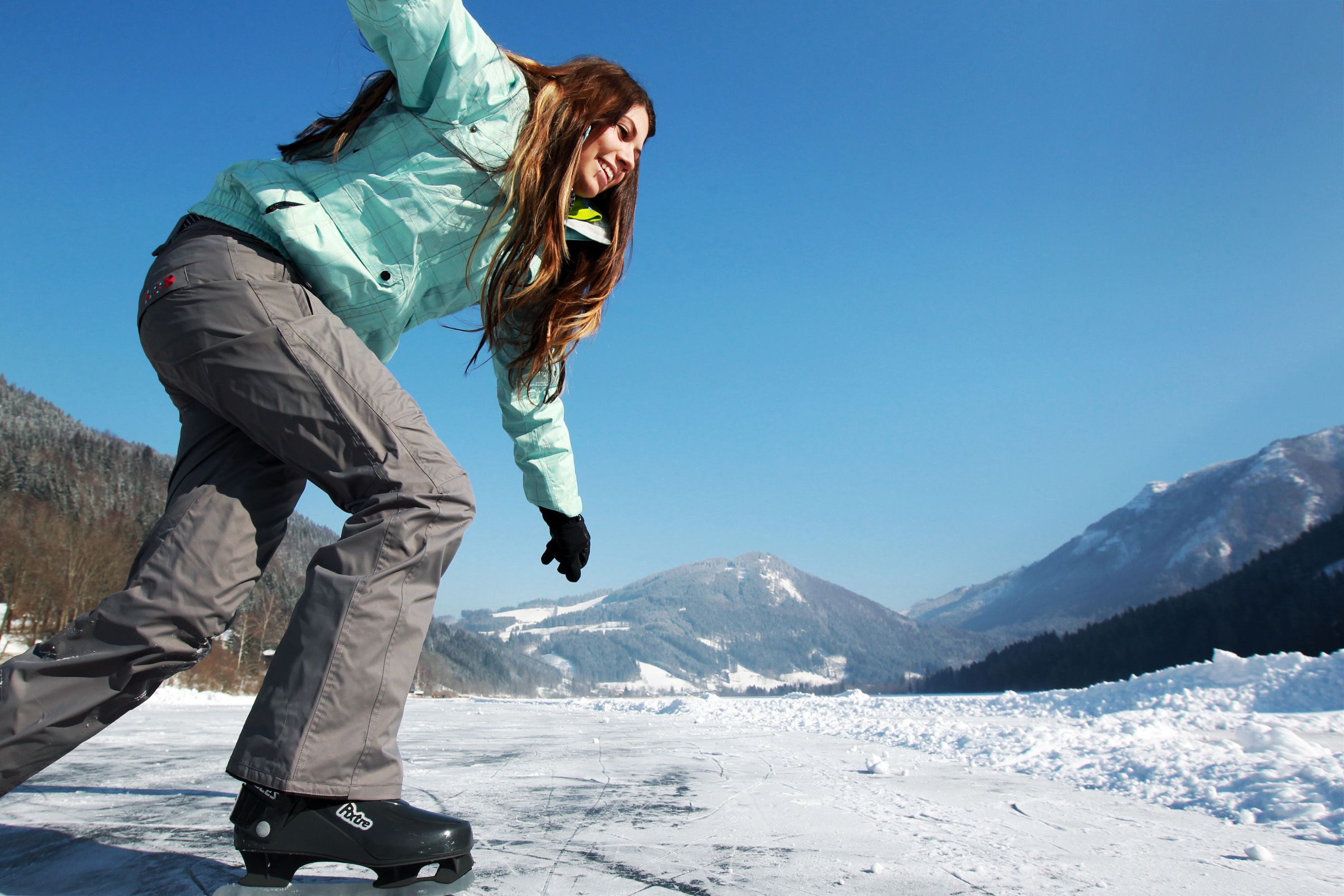 Eine Person eisläuft auf einem zugefrorenen See mit schneebedeckten Bergen im Hintergrund.
