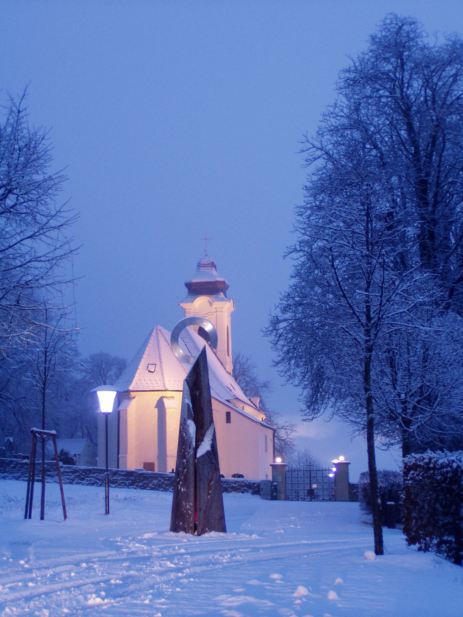 Verschneite Kirche mit beleuchtetem Turm und Skulptur im Vordergrund.