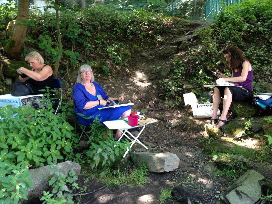 Drei Frauen sitzen im Freien und zeichnen in einem bewaldeten Gebiet.