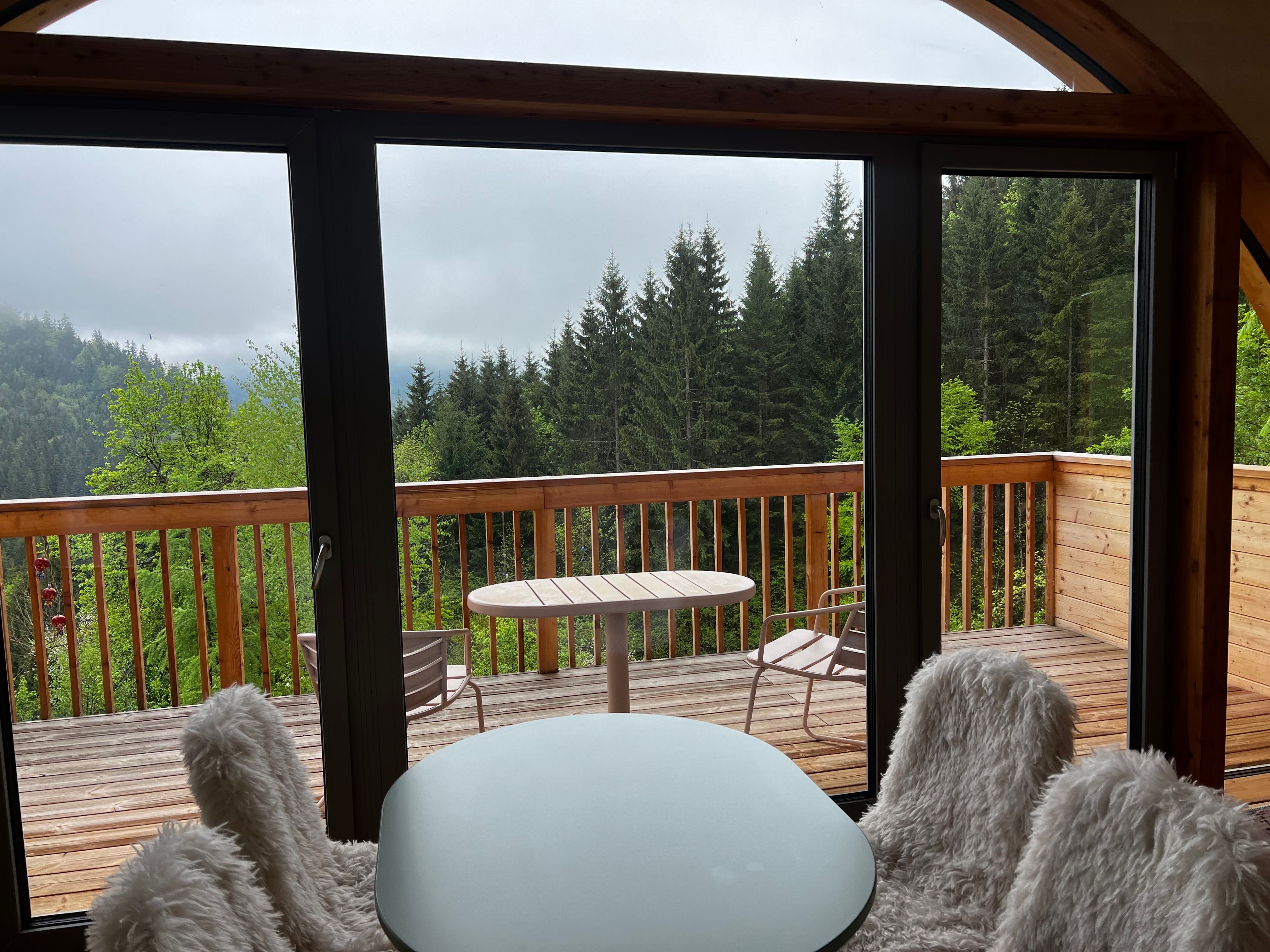 Blick von einem Innenraum auf eine Holzterrasse mit Wald im Hintergrund.