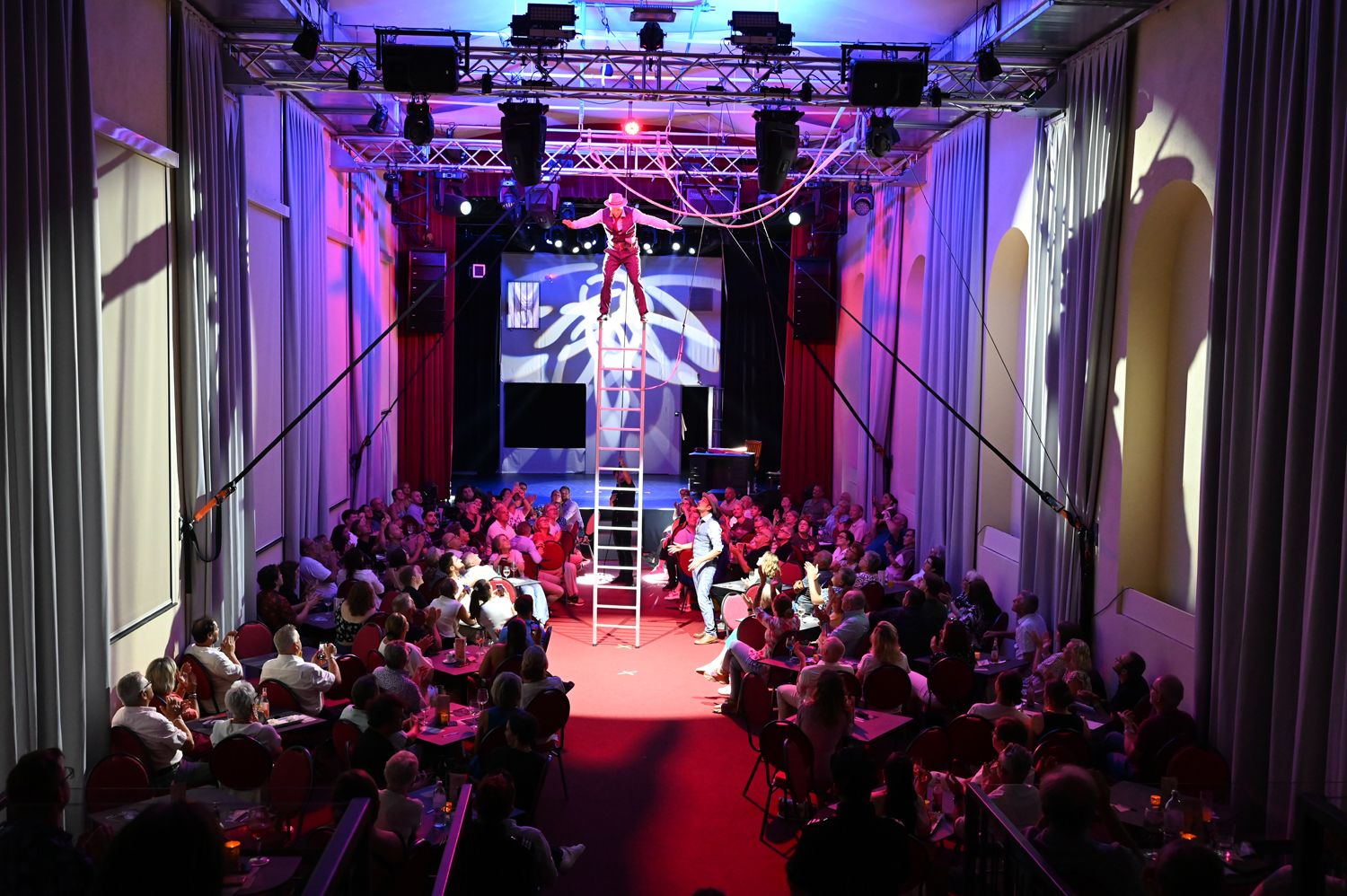 Ein Artist balanciert auf einer Leiter in einem vollbesetzten Theater mit bunter Beleuchtung.