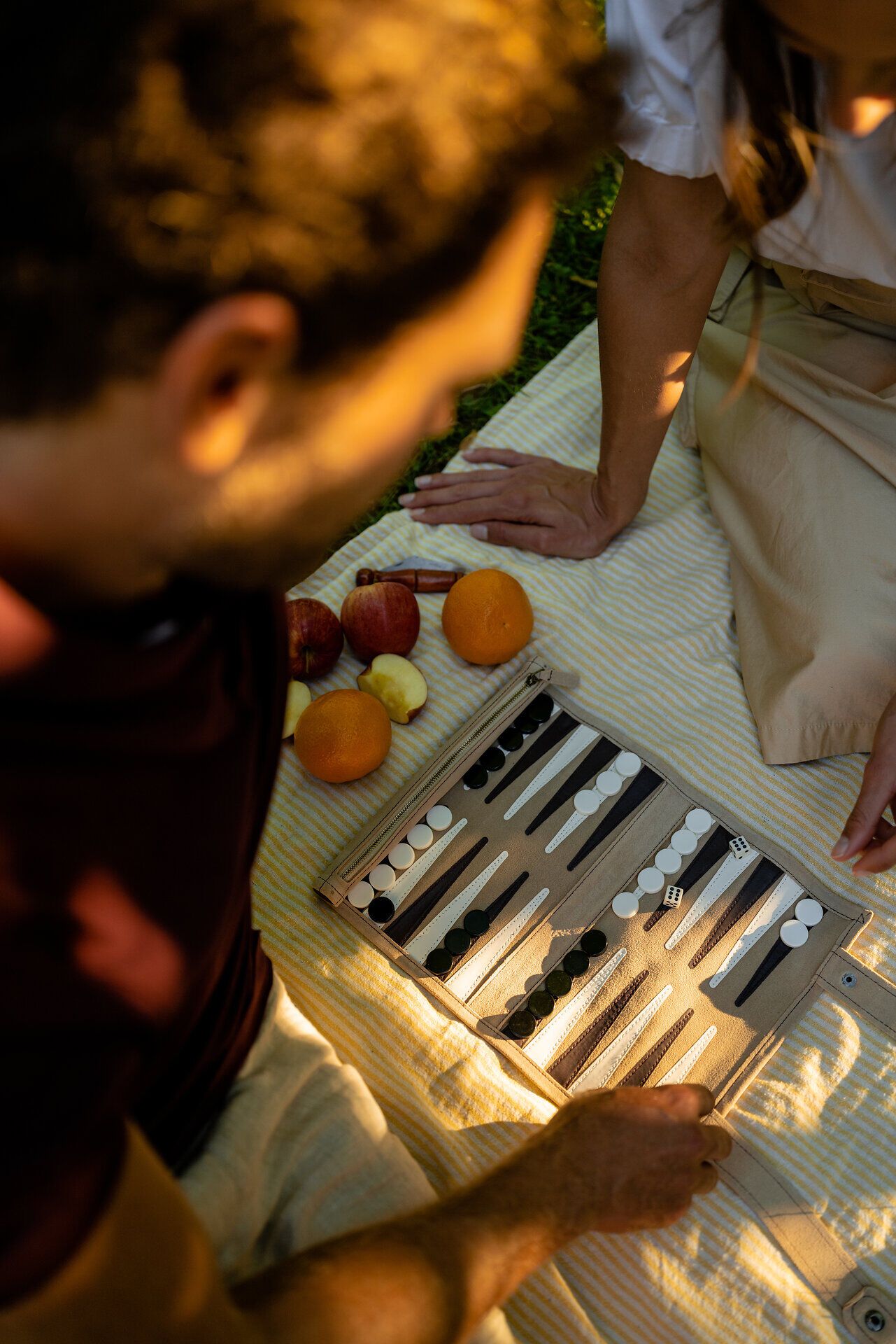 Ein entspannter Nachmittag im Freien, umgeben von saftigem Grün und dem Duft frischer Früchte. Zwei Freunde genießen ein spannendes Spiel auf einem stilvollen Backgammon-Brett, während die Sonne sanft auf die Decke scheint. Der perfekte Ort für ein Picknick und gesellige Stunden.