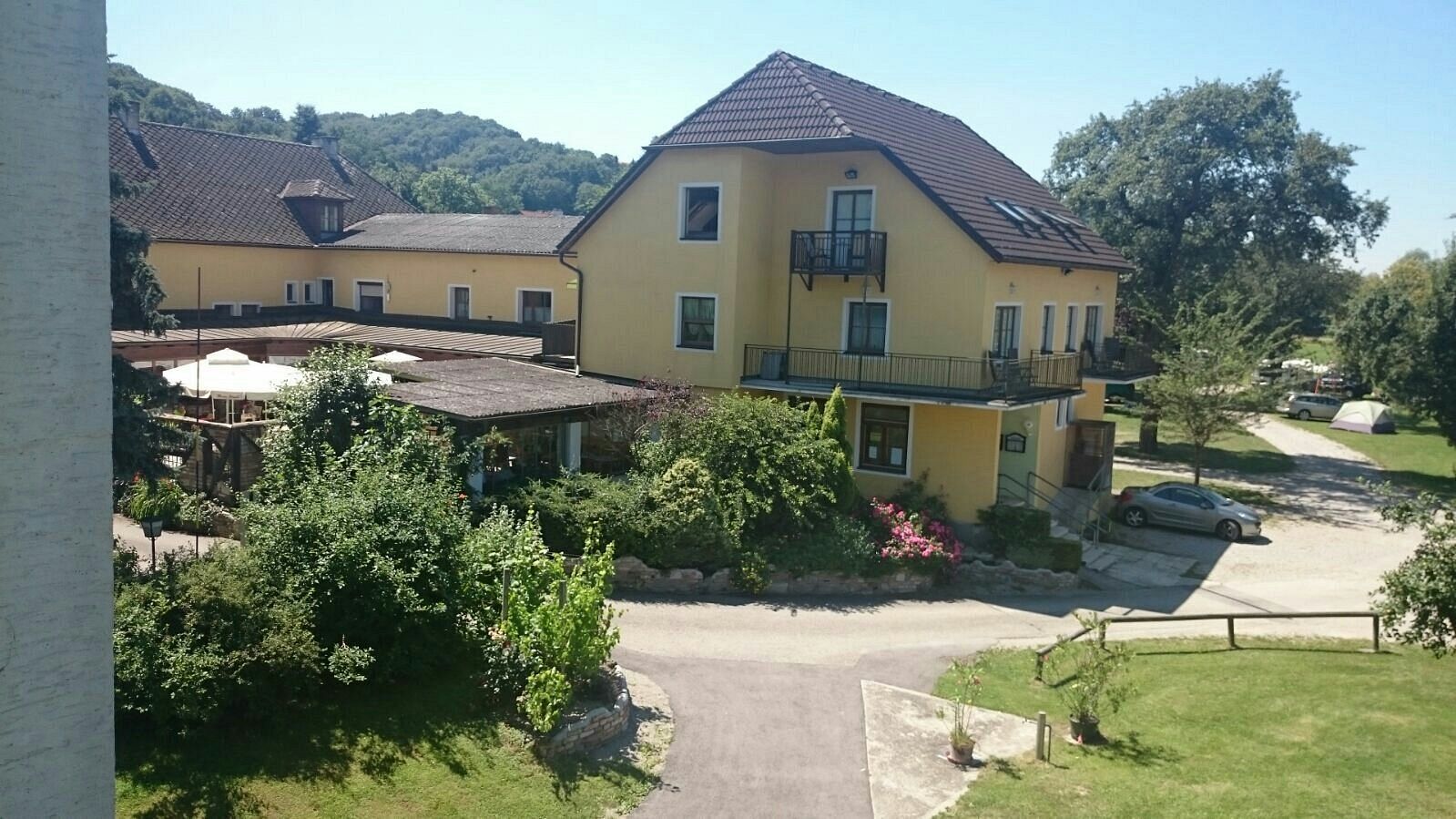Gelbes Gebäude mit Terrasse und Garten, umgeben von Bäumen und einem Auto auf dem Parkplatz.