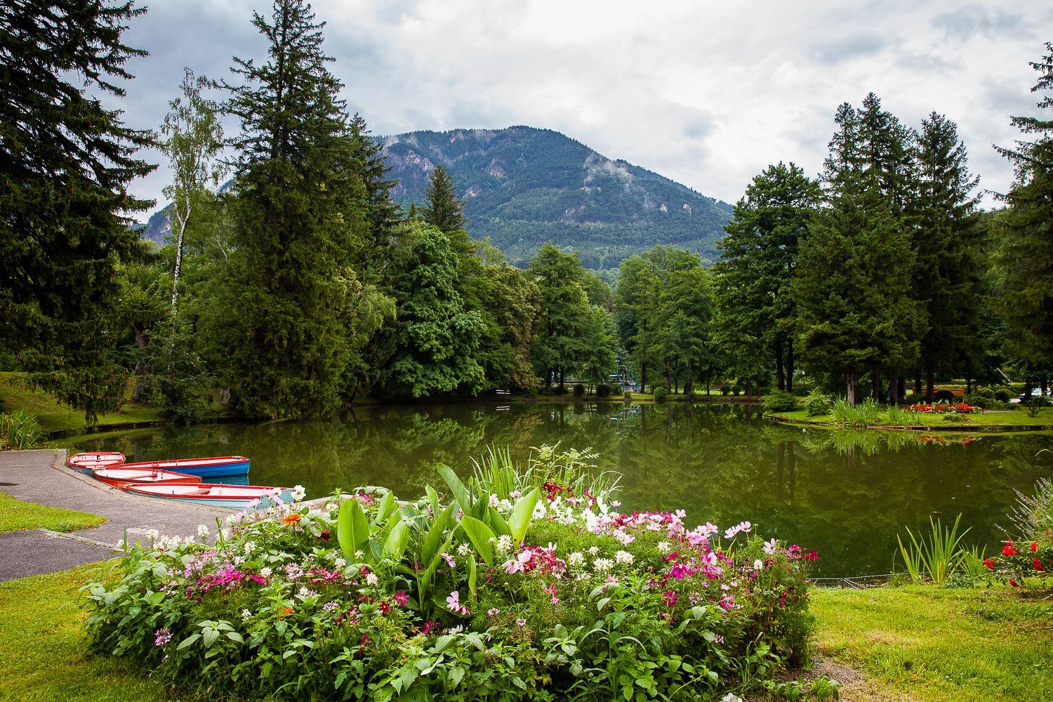 Ein malerischer Park mit einem Teich, umgeben von Bäumen und Blumen, im Hintergrund Berge.