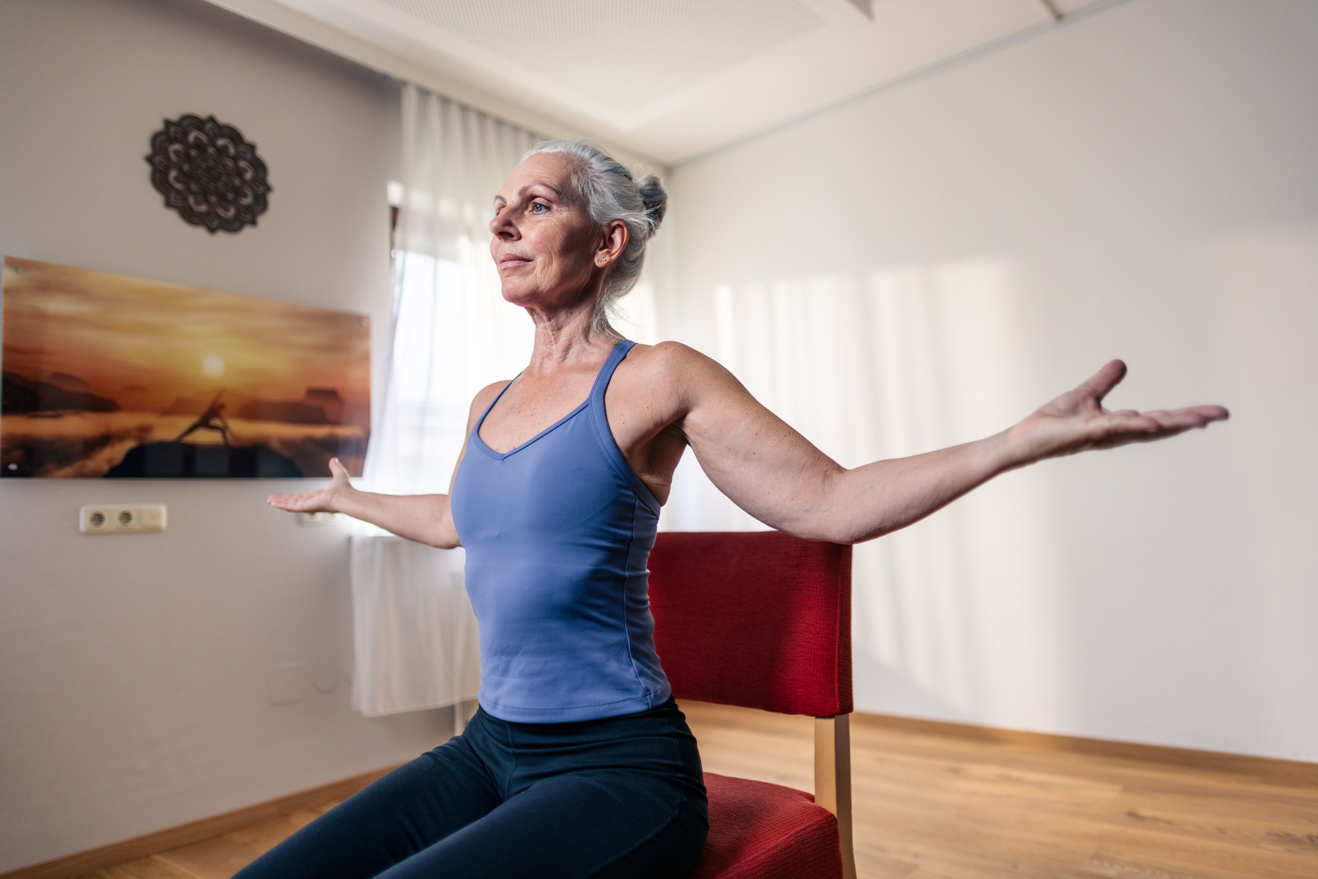 Eine ältere Frau macht Yoga auf einem Stuhl in einem hellen Raum.