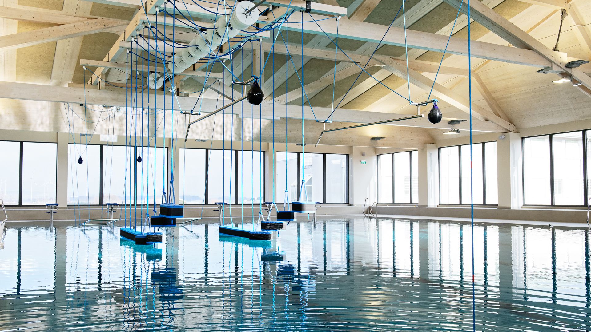 Innenansicht eines modernen Schwimmbads mit hängenden blauen Seilen und Plattformen über dem Wasser.