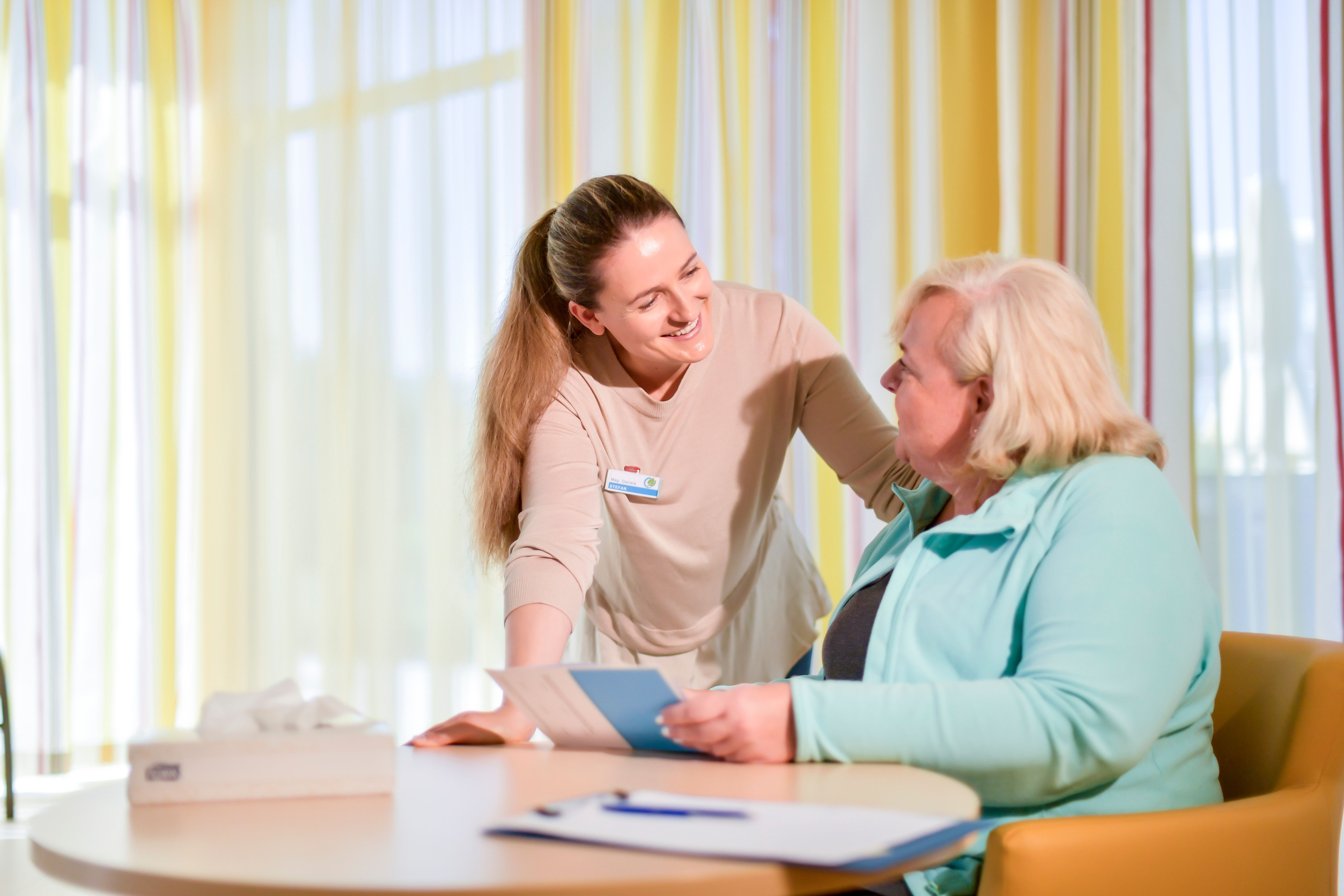 Eine Frau spricht lächelnd mit einer anderen Frau an einem Tisch mit Dokumenten.