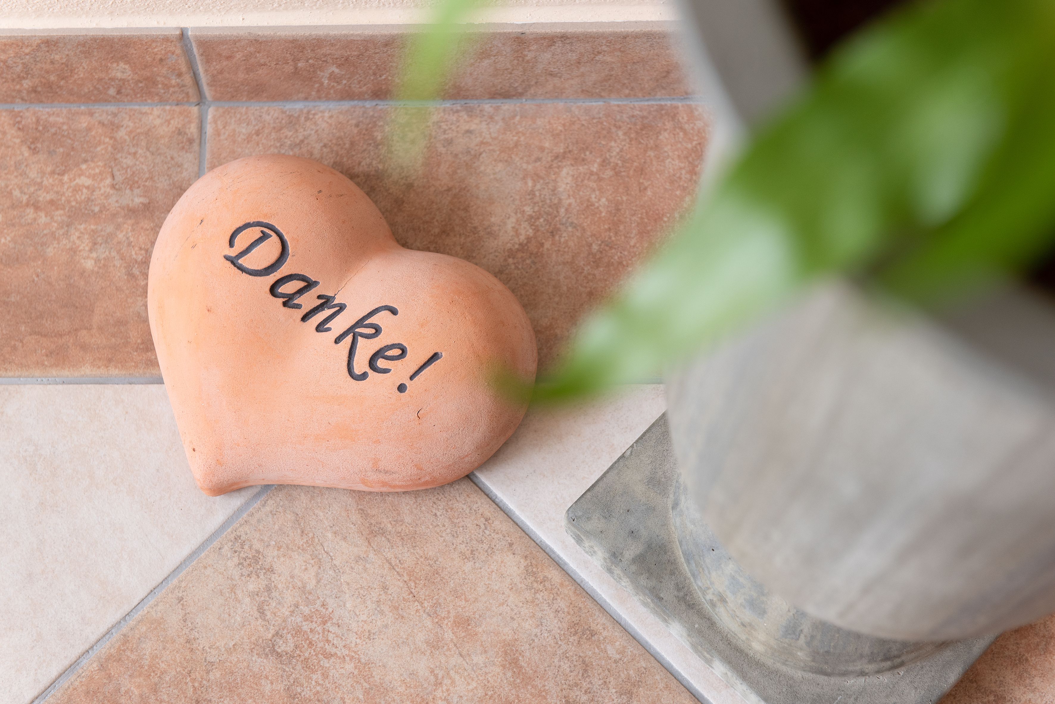 Herzförmiger Stein mit der Aufschrift 'Danke!' auf Fliesenboden neben Pflanzentopf.