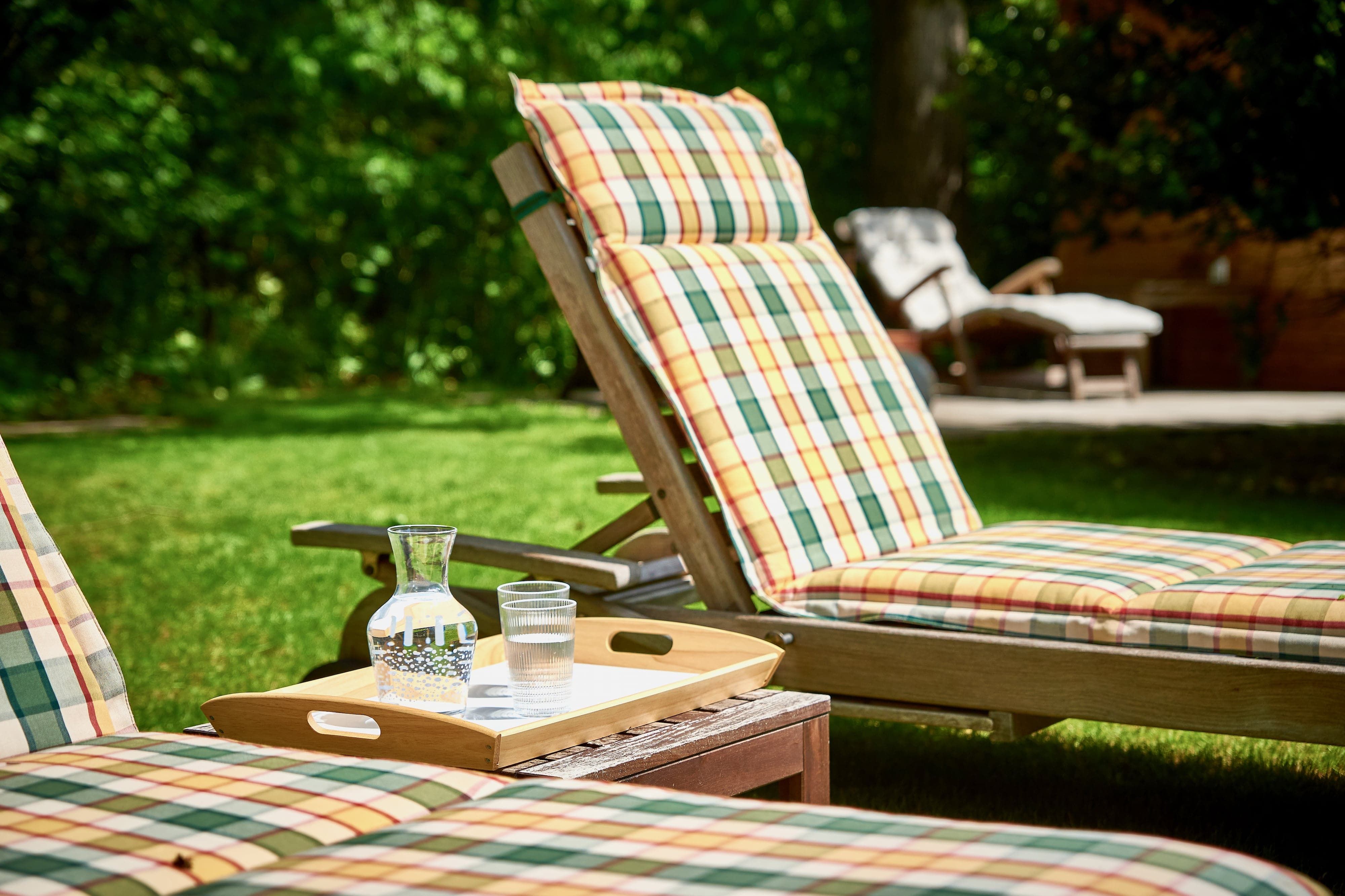 Sonnenliegen mit karierten Auflagen und einem Tablett mit Wasser im Garten.