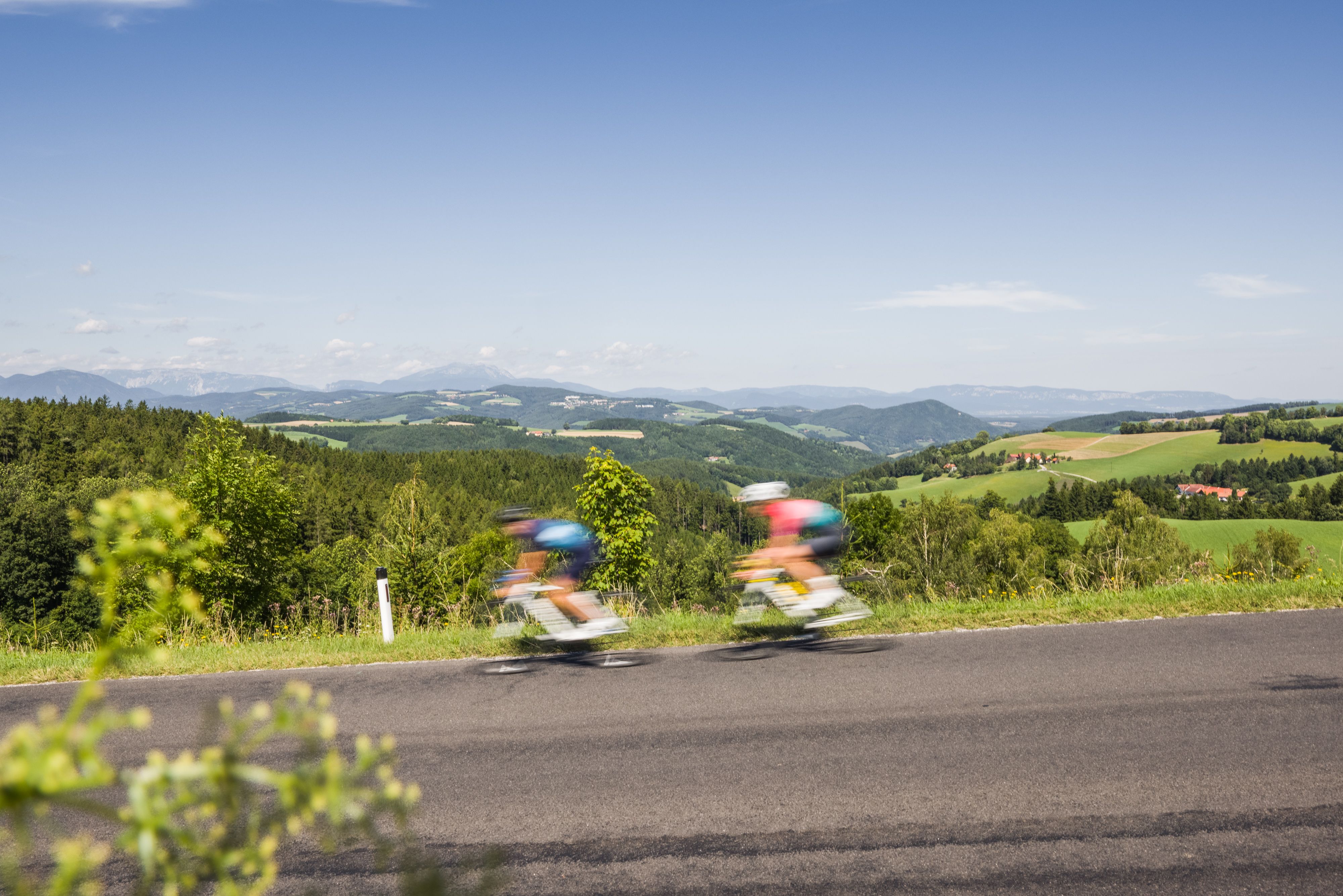 Zwei Personen am Rennrad fahren schnell auf einer Straße in einer hügeligen Landschaft.