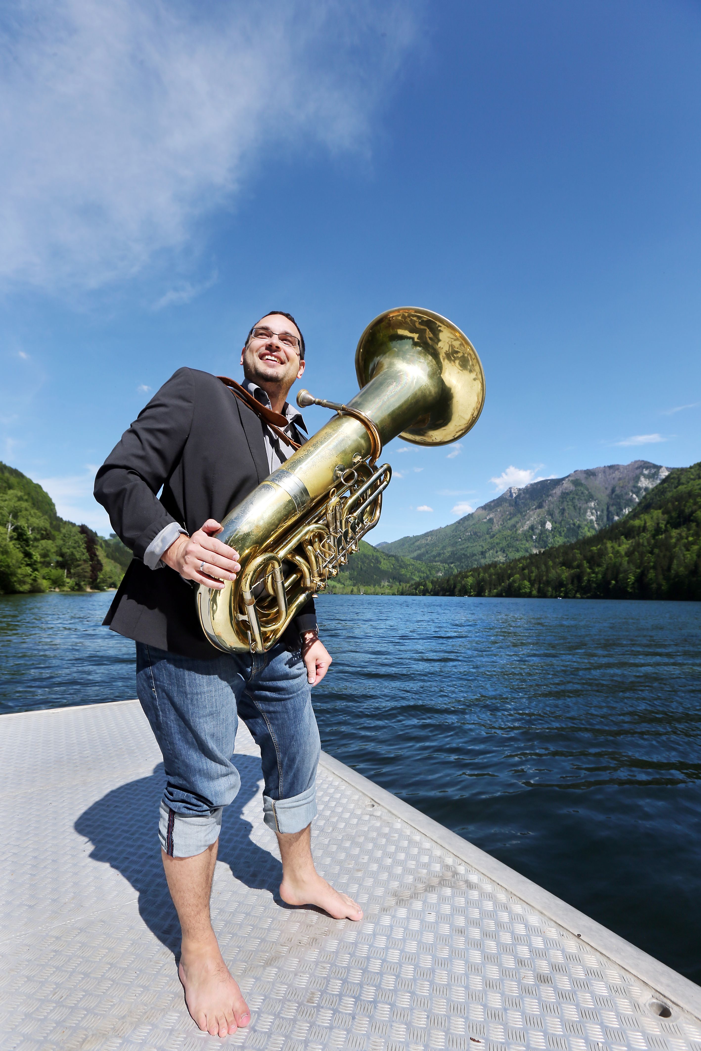 Ein Mann mit einer Tuba steht barfuß auf einem Steg an einem See, umgeben von Bergen und blauem Himmel.