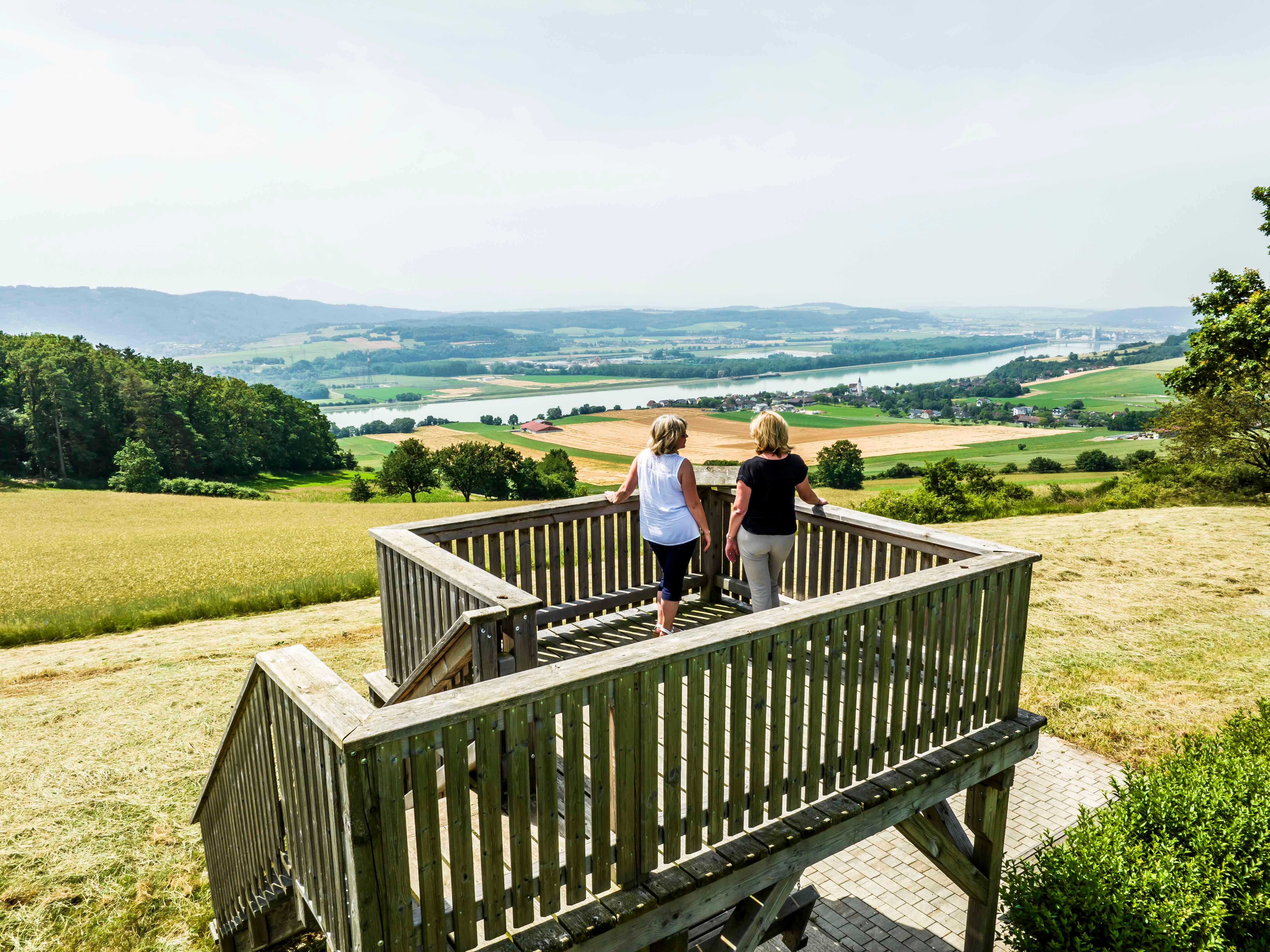Zwei Personen stehen auf einer hölzernen Aussichtsplattform mit Blick auf eine weite Landschaft und einen Fluss.