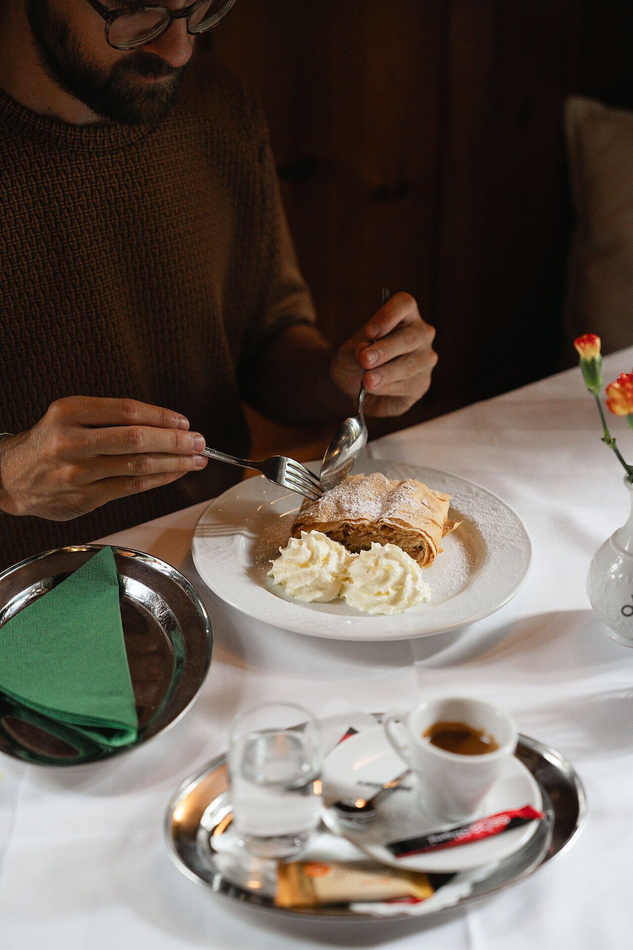Ein köstlicher Apfelstrudel mit Schlagobers steht neben einer Tasse frisch zubereitetem Kaffee auf einem Holztisch mit weißem Tischtuch. Ideal für eine genussvolle Auszeit in rustikaler Umgebung.