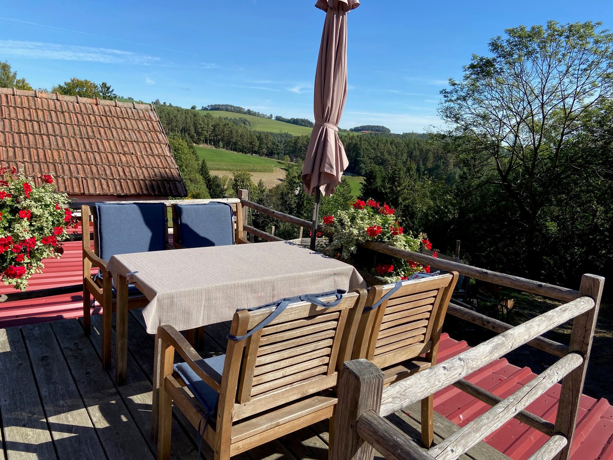 Kleine Terrasse mit Holzmöbeln, Sonnenschirm und Blumenkästen, umgeben von grüner Landschaft.