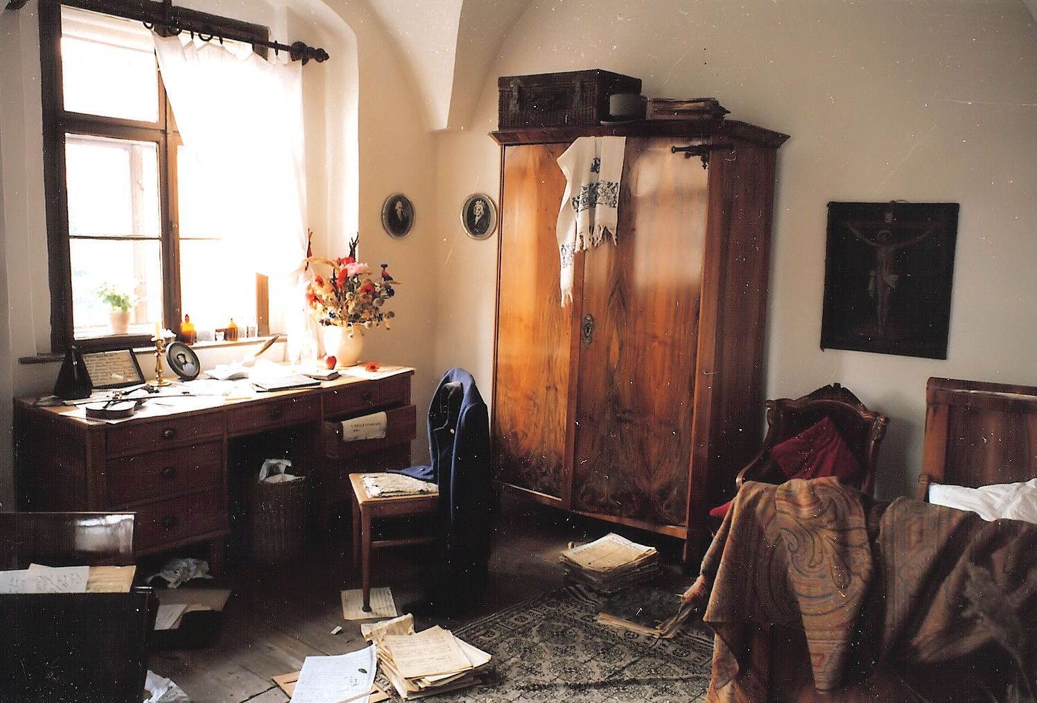 Ein historischer Raum mit einem Holztisch, Notenblättern, einem Schrank und einem Bett. Der Raum wirkt unordentlich und altmodisch.