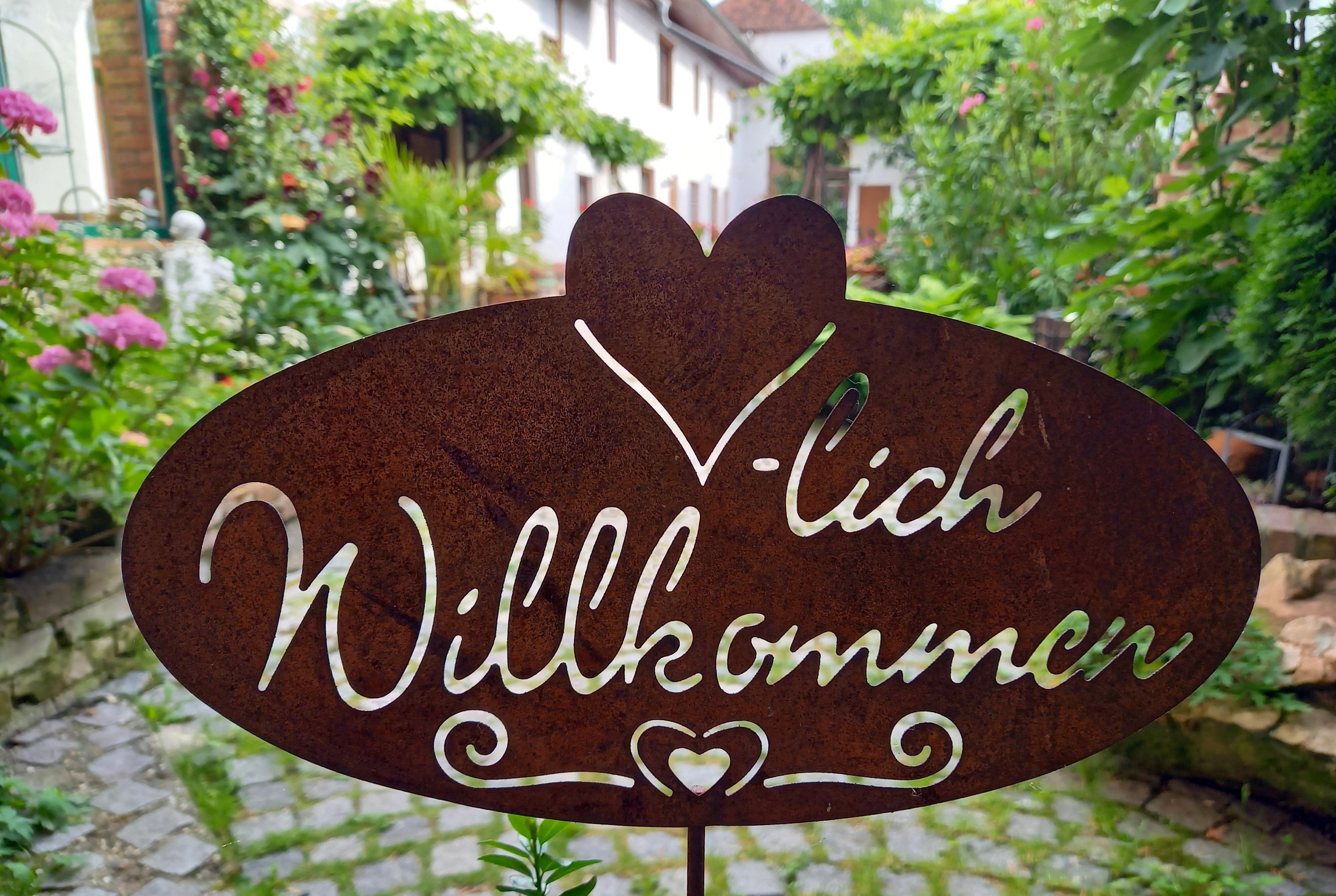 Metallschild mit der Aufschrift 'Herzlich Willkommen' in einem Garten.