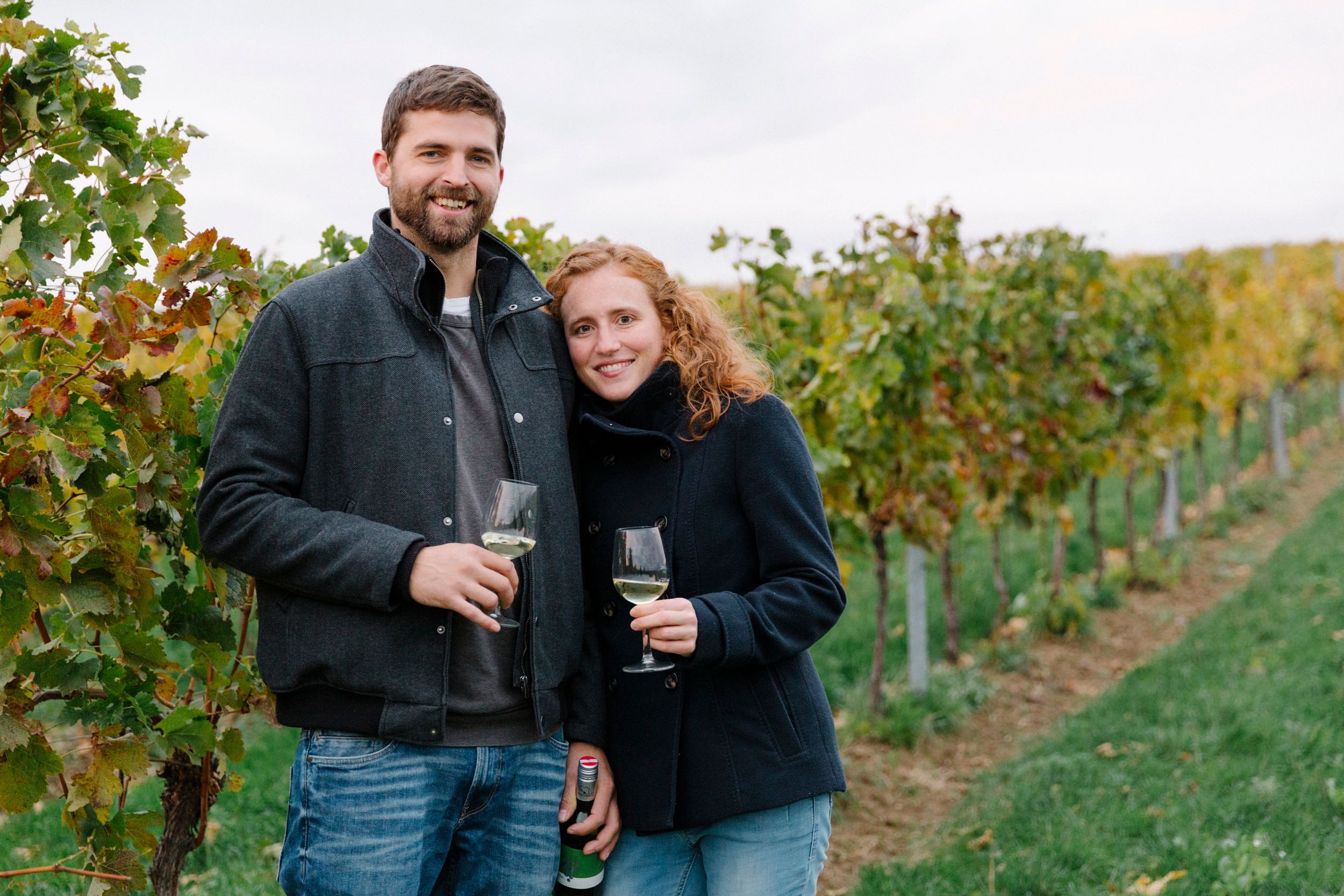 Ein Paar steht in einem Weinberg und hält Weingläser in der Hand.