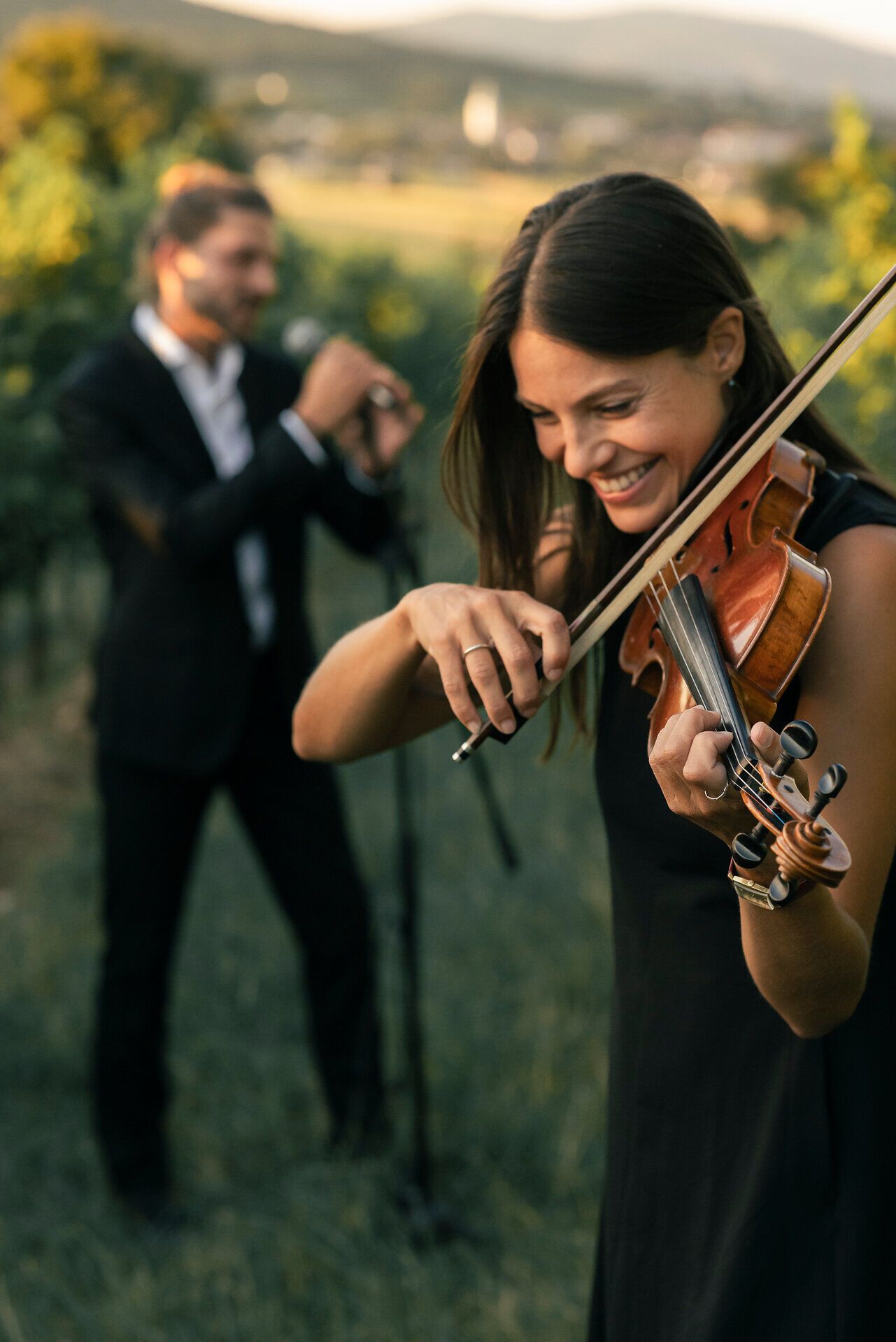 Eine Frau spielt draußen fröhlich Geige, während ein Mann im Hintergrund eins Mikrofon singt. Beide sind elegant gekleidet, und die Umgebung scheint ein weinbergähnliches Setting zu sein.
