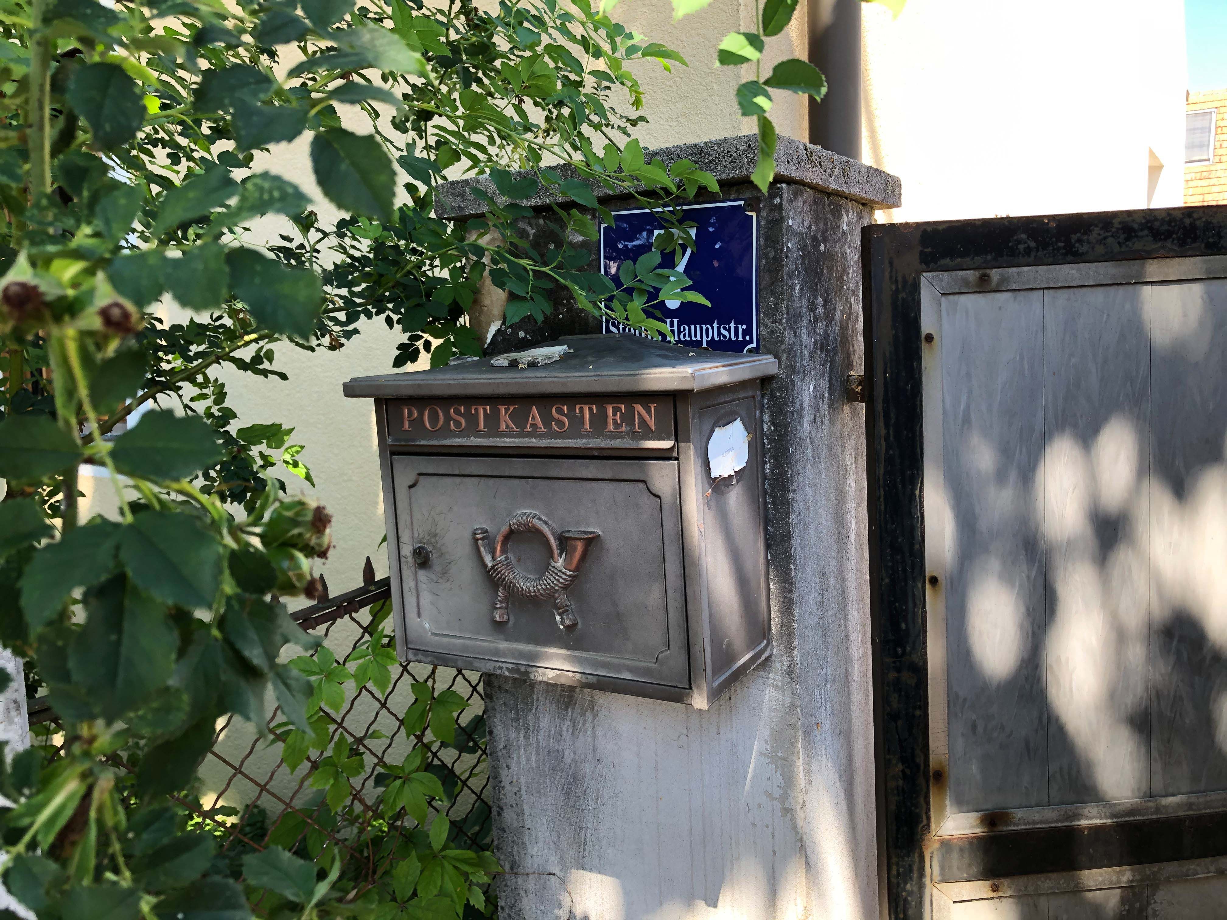 Ein alter Briefkasten mit der Aufschrift 'Postkasten' an einer Mauer neben einem Tor, umgeben von Pflanzen.