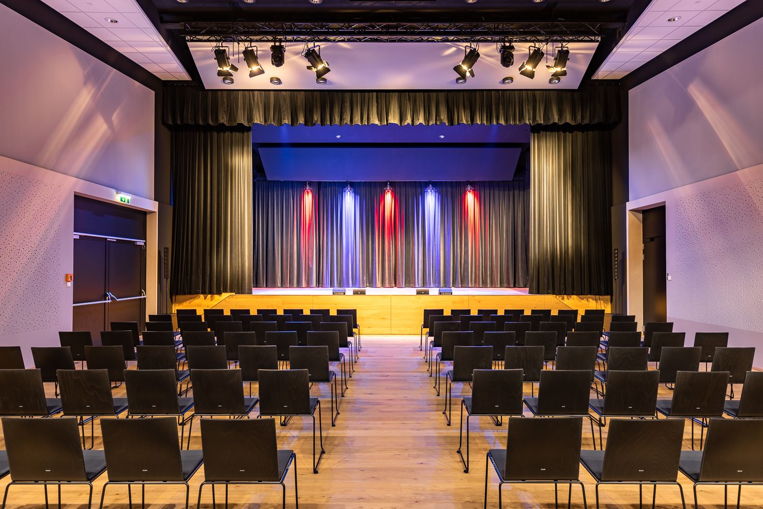 Ein leerer Veranstaltungssaal mit Stuhlreihen und einer Bühne mit Vorhängen und bunter Beleuchtung.