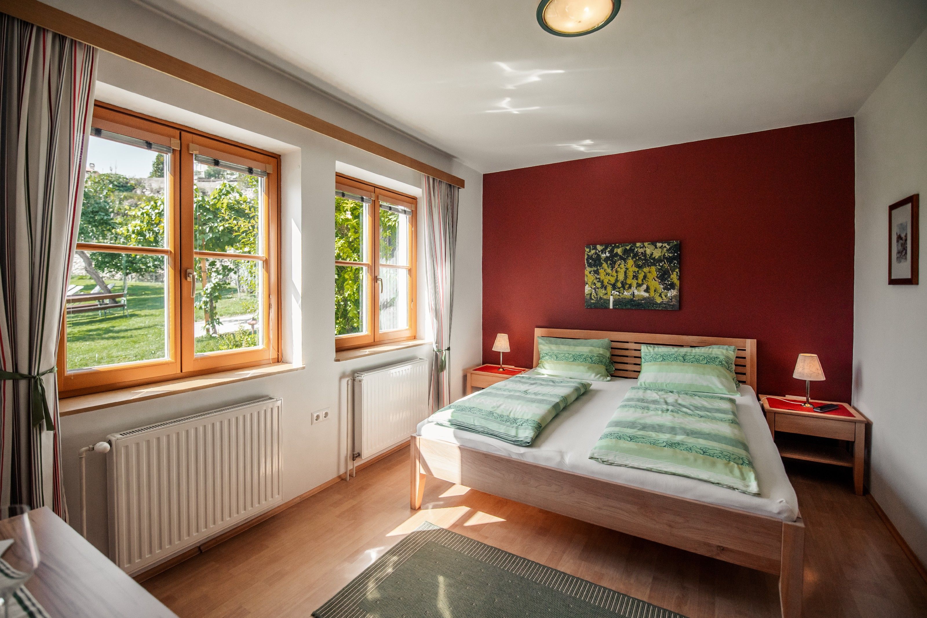 Helles Schlafzimmer mit Doppelbett, roten Wänden und großen Fenstern mit Blick ins Grüne.