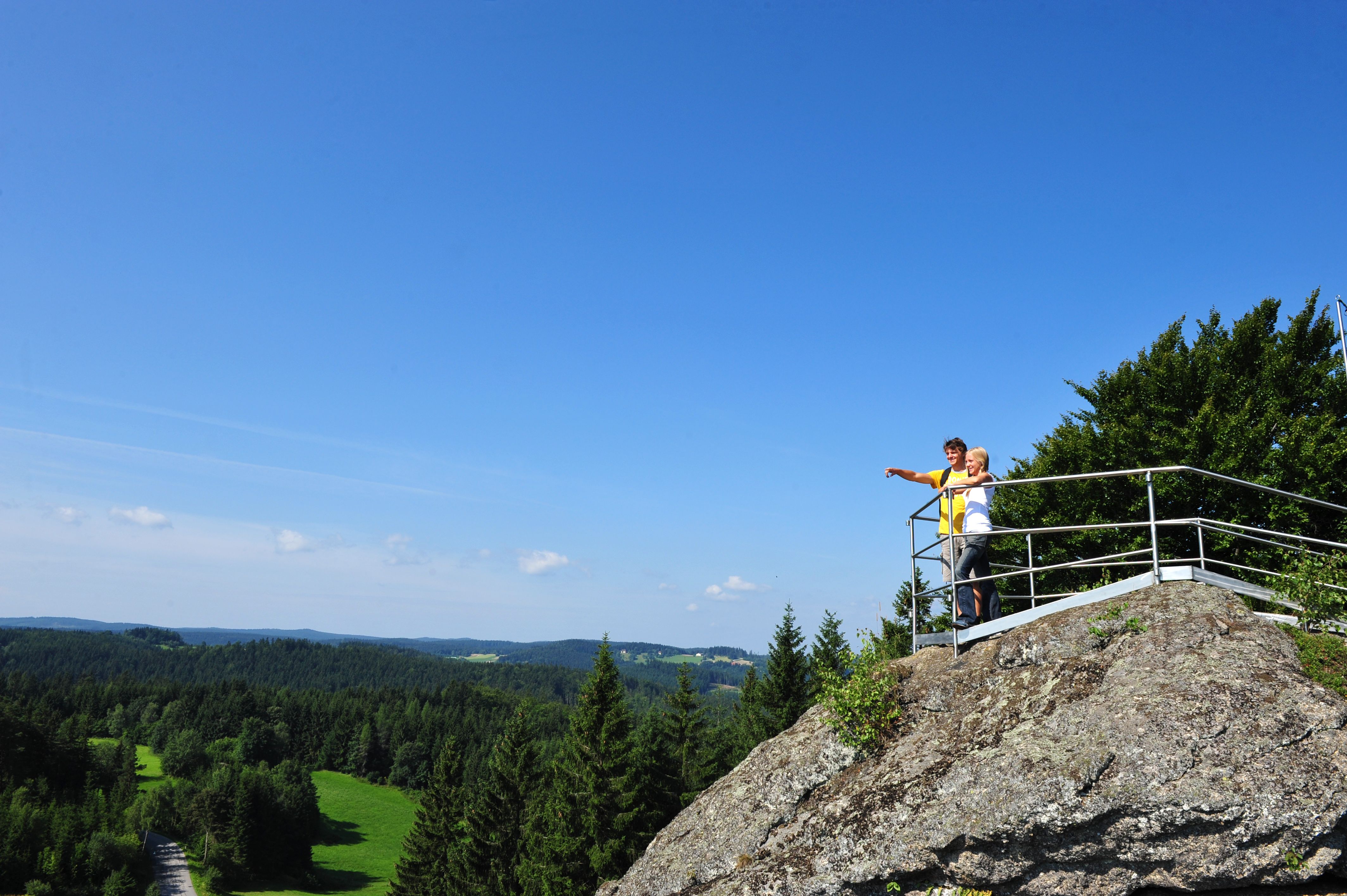 Zwei Personen stehen auf einem Aussichtspunkt auf einem Felsen und blicken in die Ferne über eine bewaldete Landschaft.