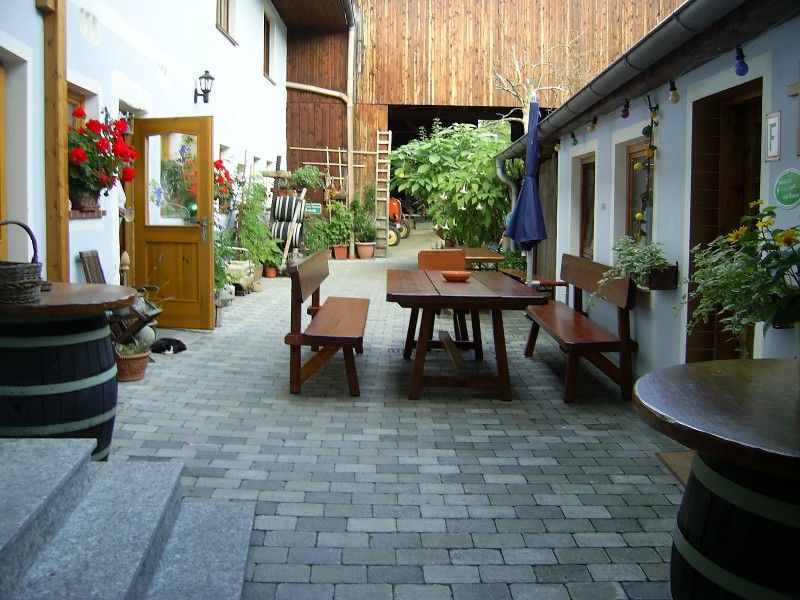 Innenhof eines Bauernhofs mit Holztischen und Bänken, umgeben von Pflanzen und Blumen.