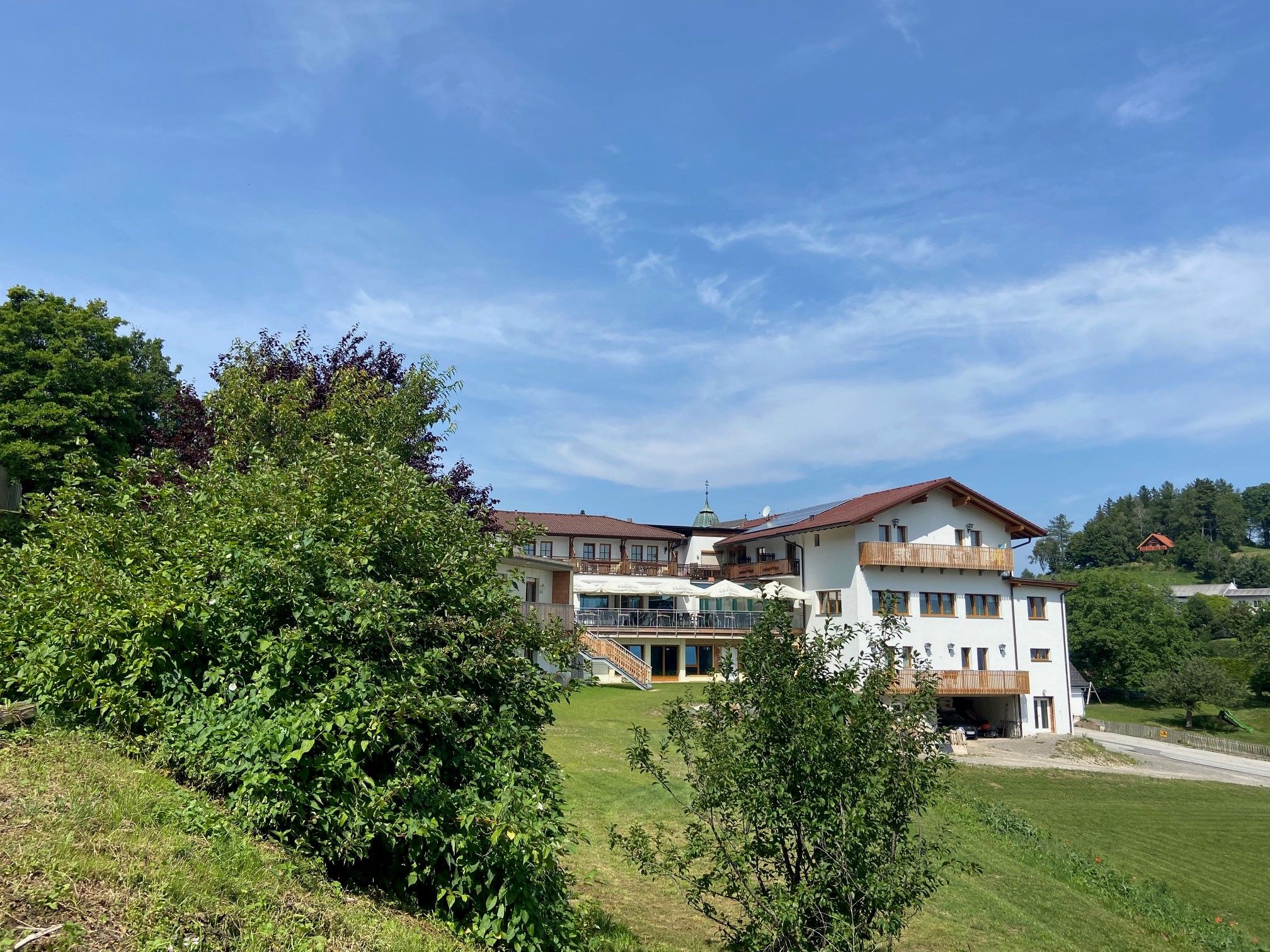 Großes Gebäude mit roten Dächern und Balkonen, umgeben von Bäumen und Wiesen unter blauem Himmel.