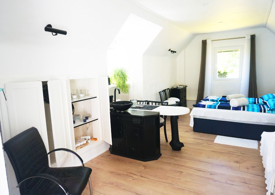 Innenansicht eines hellen Ferienhauses mit Bett, Tisch, Stühlen und Küchenzeile.