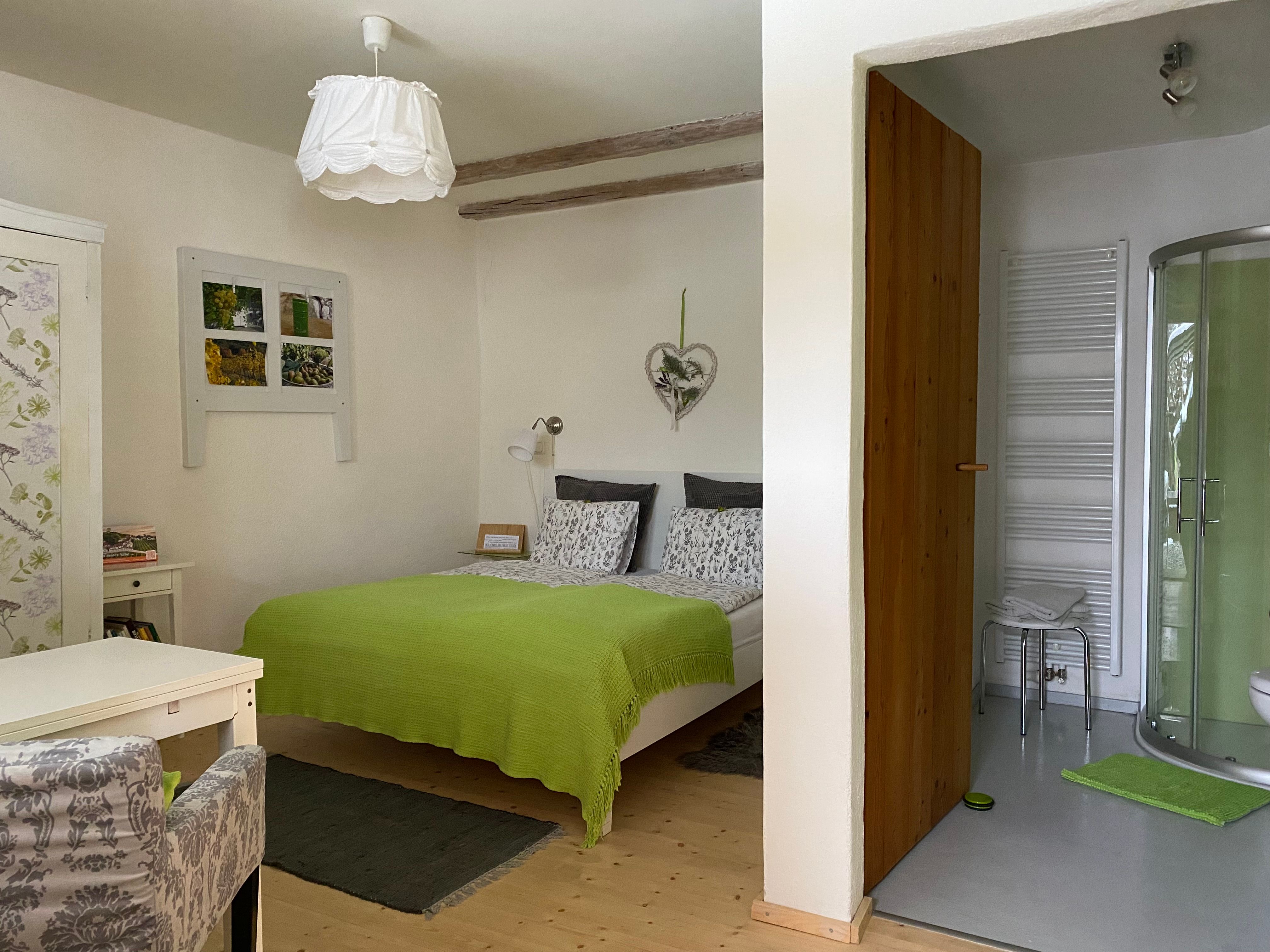 Gemütliches Doppelzimmer mit grünem Bettüberwurf, kleinem Schreibtisch und angrenzendem Badezimmer mit Dusche.