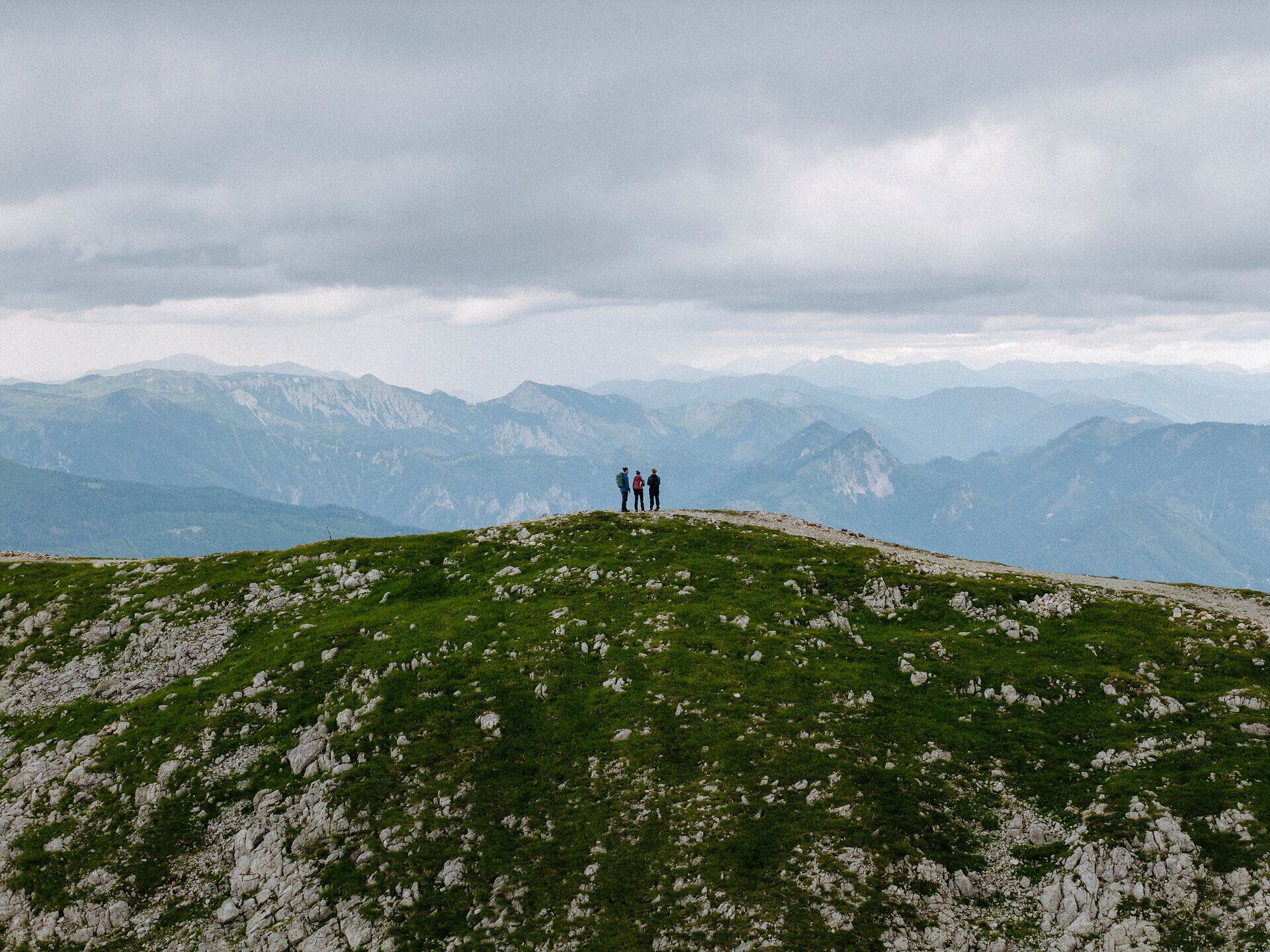 Drei wandernde Personen stehen auf einem Wanderweg entlang eines Kamms, dahinter die Berglandschaft.