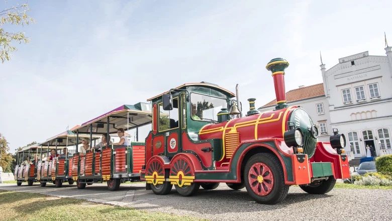 Ein bunter Touristenzug mit mehreren Waggons fährt an einem weißen Gebäude vorbei.