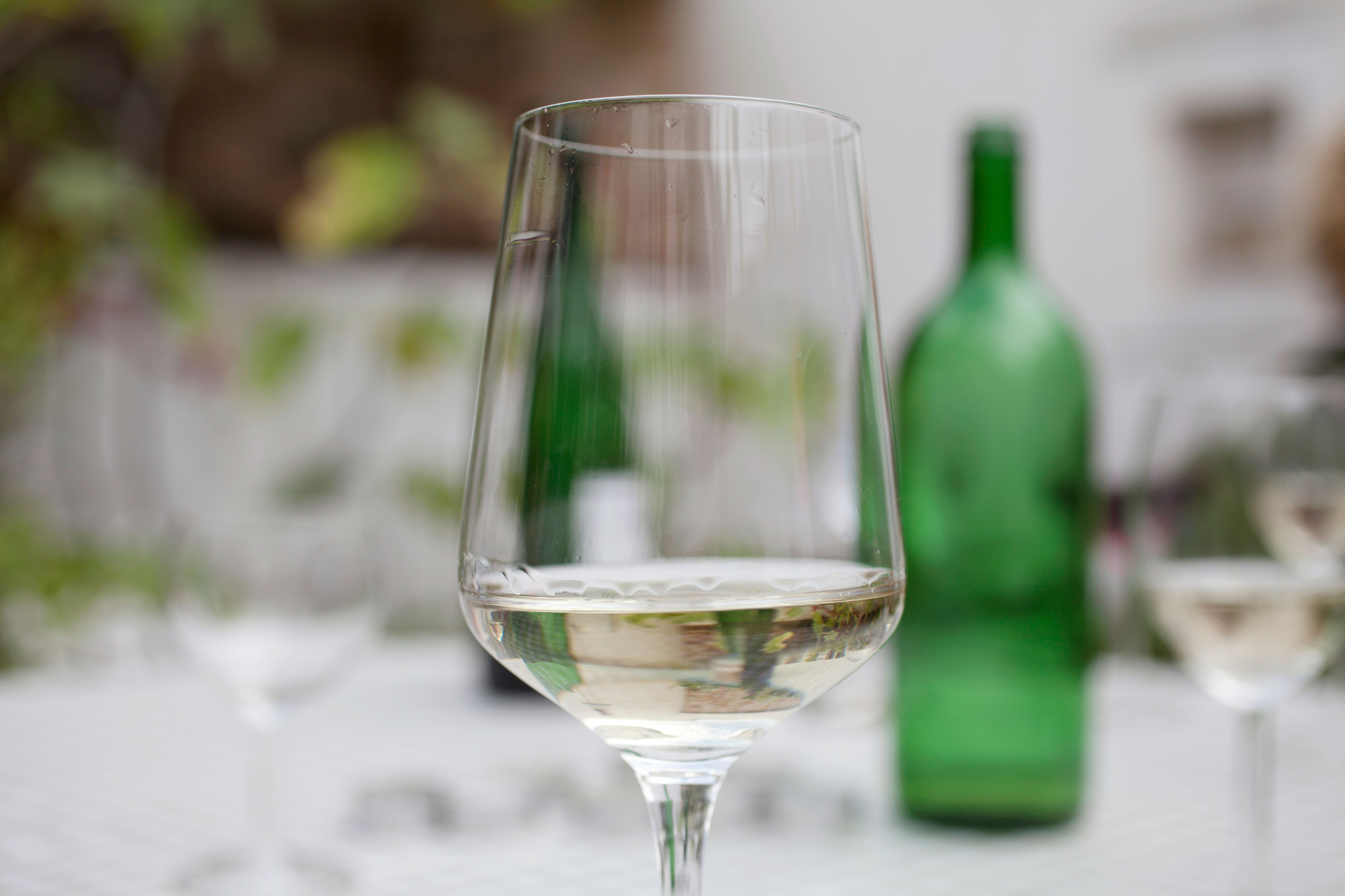 Ein Weinglas mit Weißwein im Vordergrund, unscharfe grüne Weinflasche im Hintergrund.